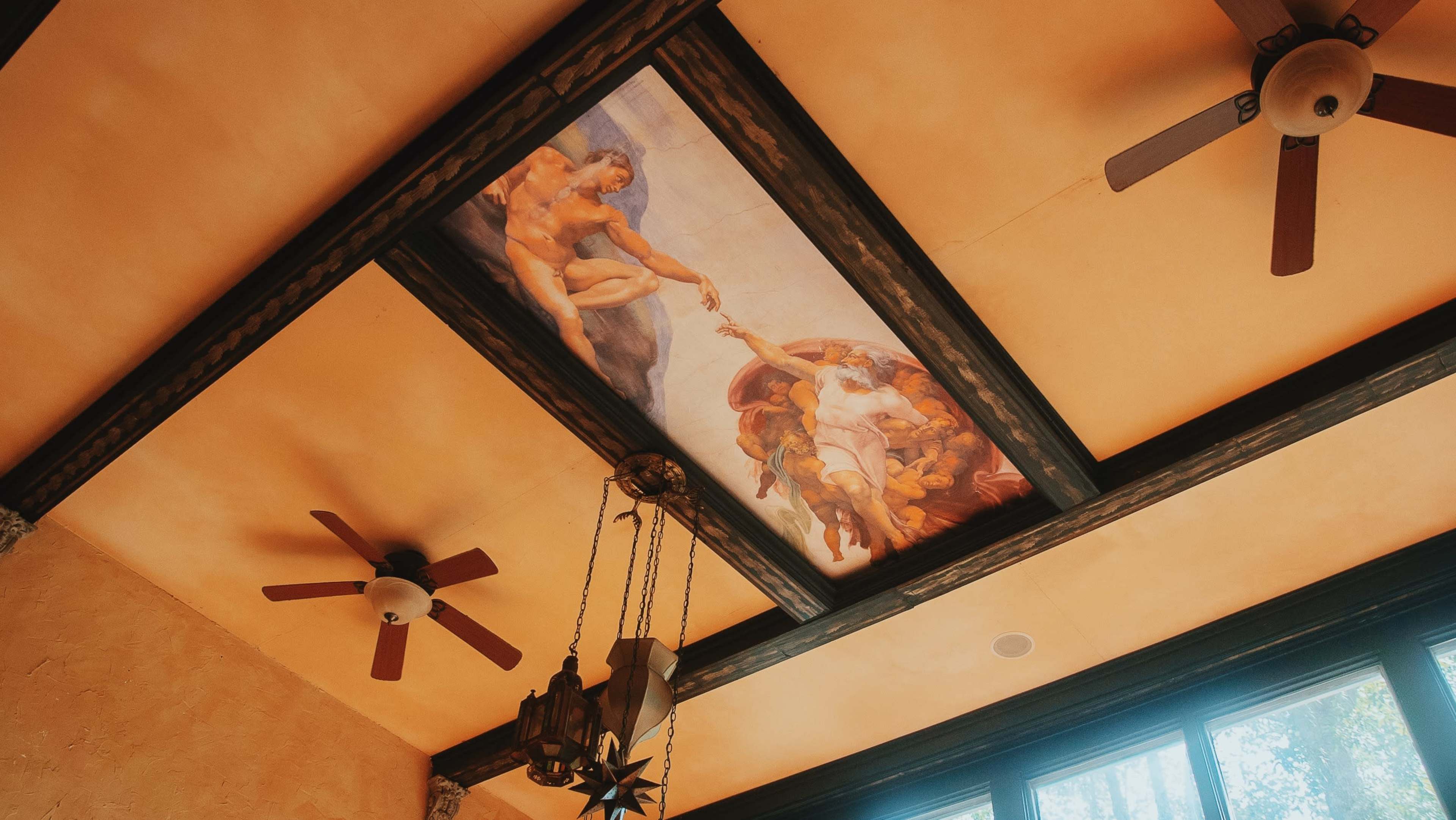 The ceiling features a painted scene resembling a classical artwork, framed by wooden beams and accompanied by ceiling fans.