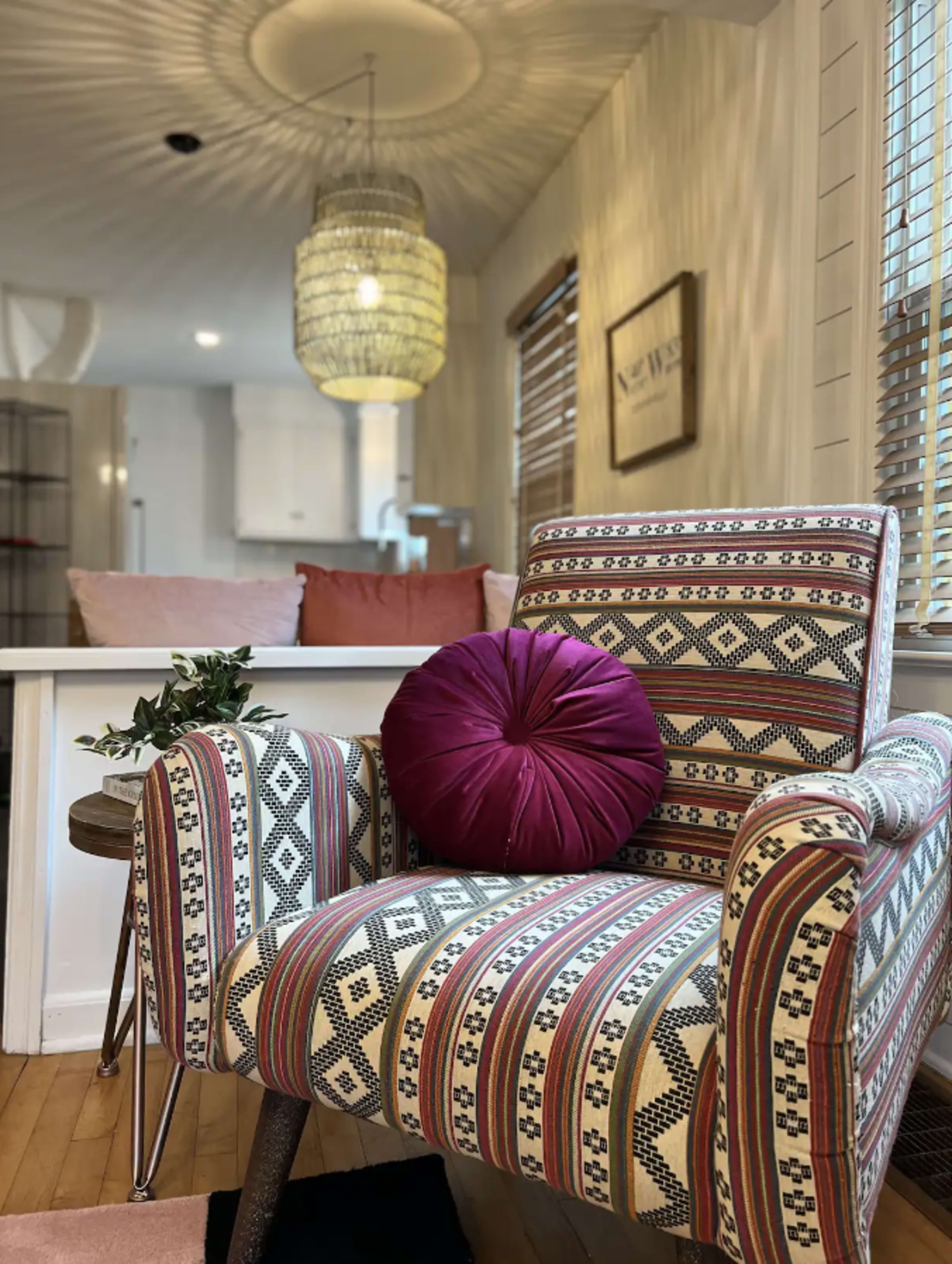 A cozy interior space featuring a patterned armchair with a decorative pillow, set against a backdrop of wooden floors and a bright, airy kitchen area.