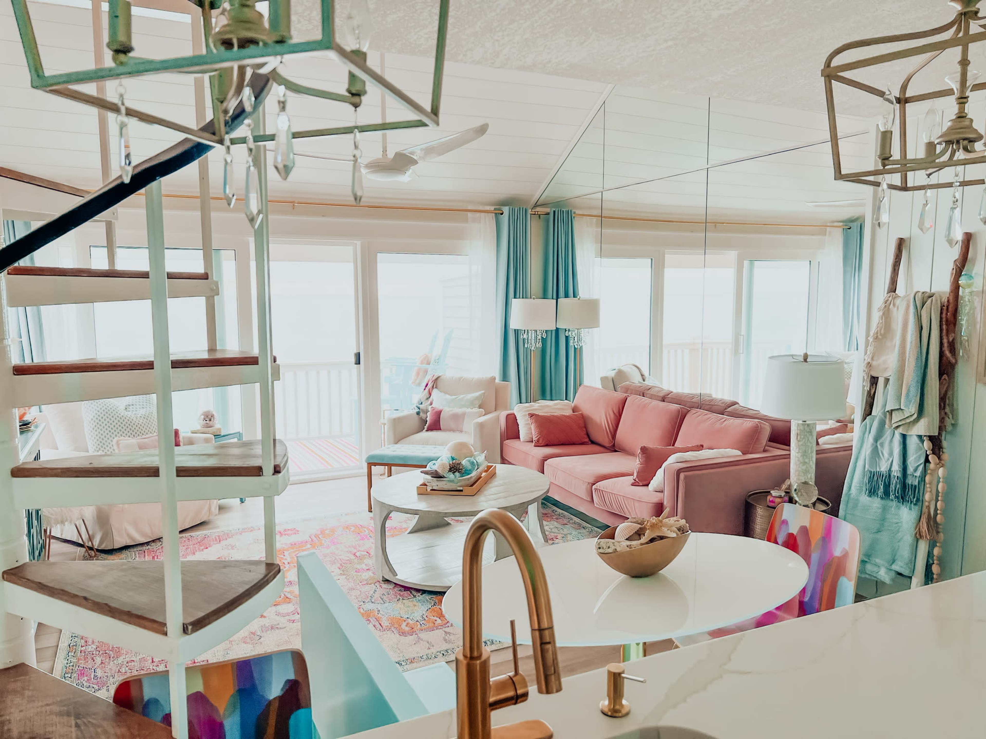 A bright, airy living space featuring a pink couch, a coffee table, and a spiral staircase leading to an upper level with large windows offering a view.
