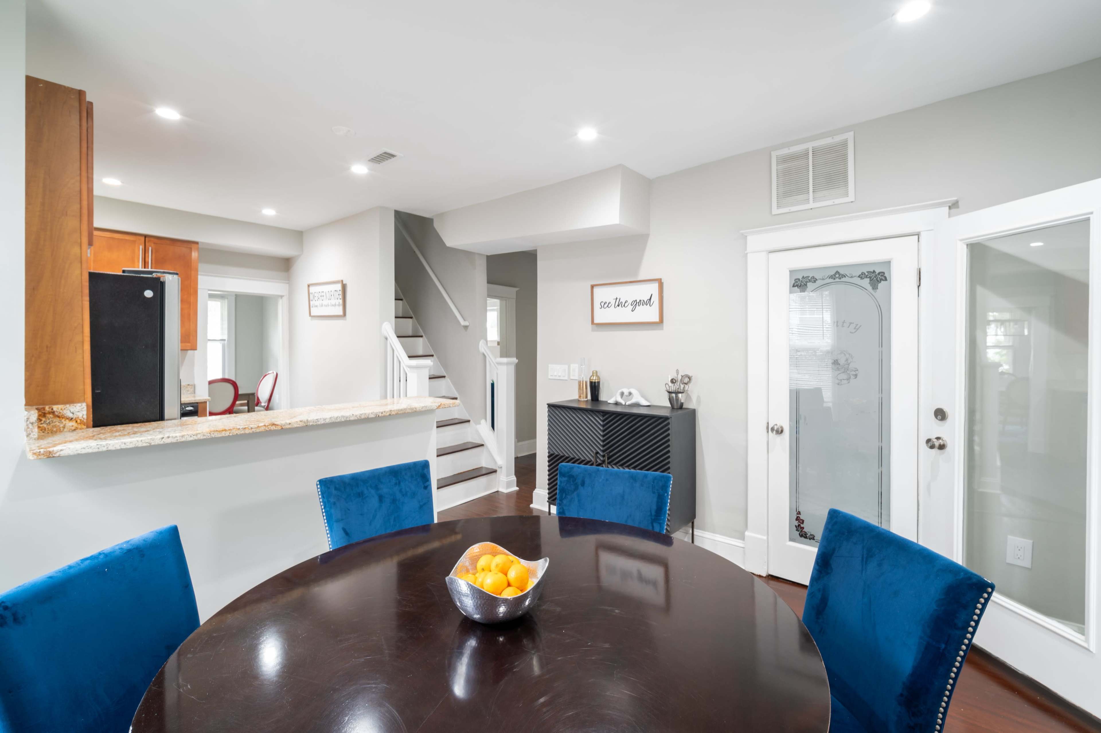 A modern dining area with a round table, blue upholstered chairs, and a bowl of oranges, adjacent to a kitchen and a staircase.