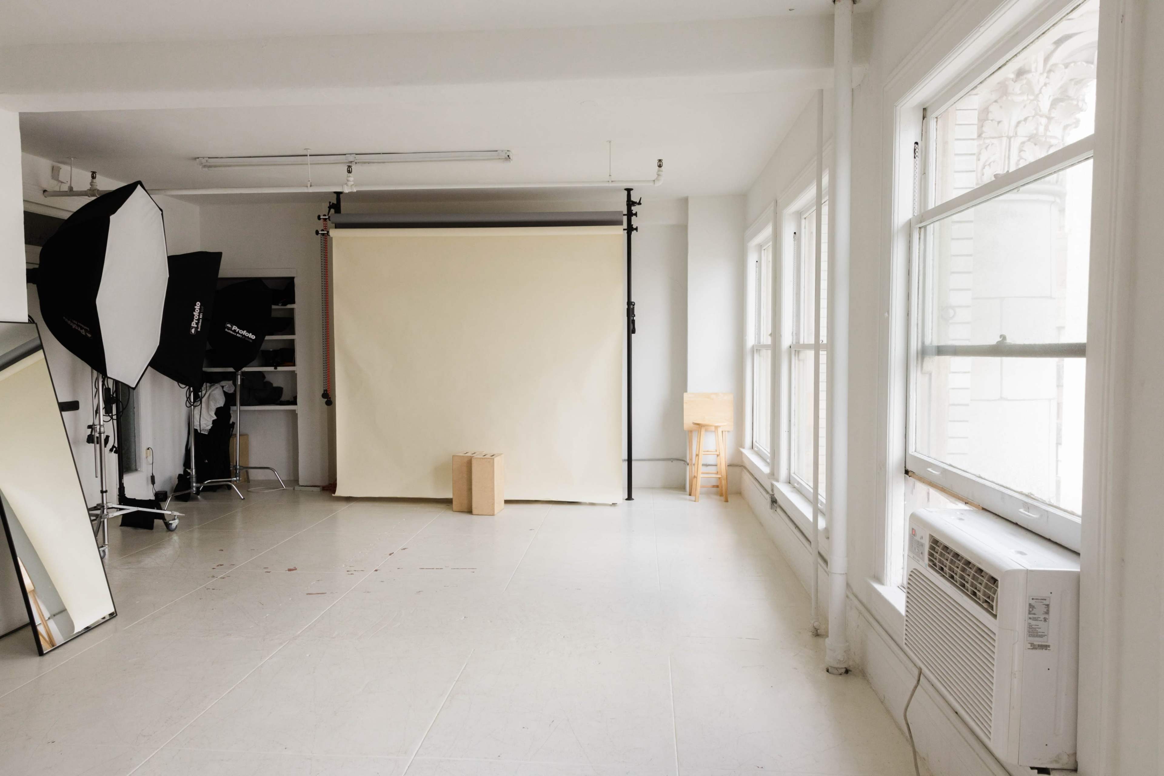 A photography studio with a light backdrop, several lighting equipment, a mirror, and large windows.