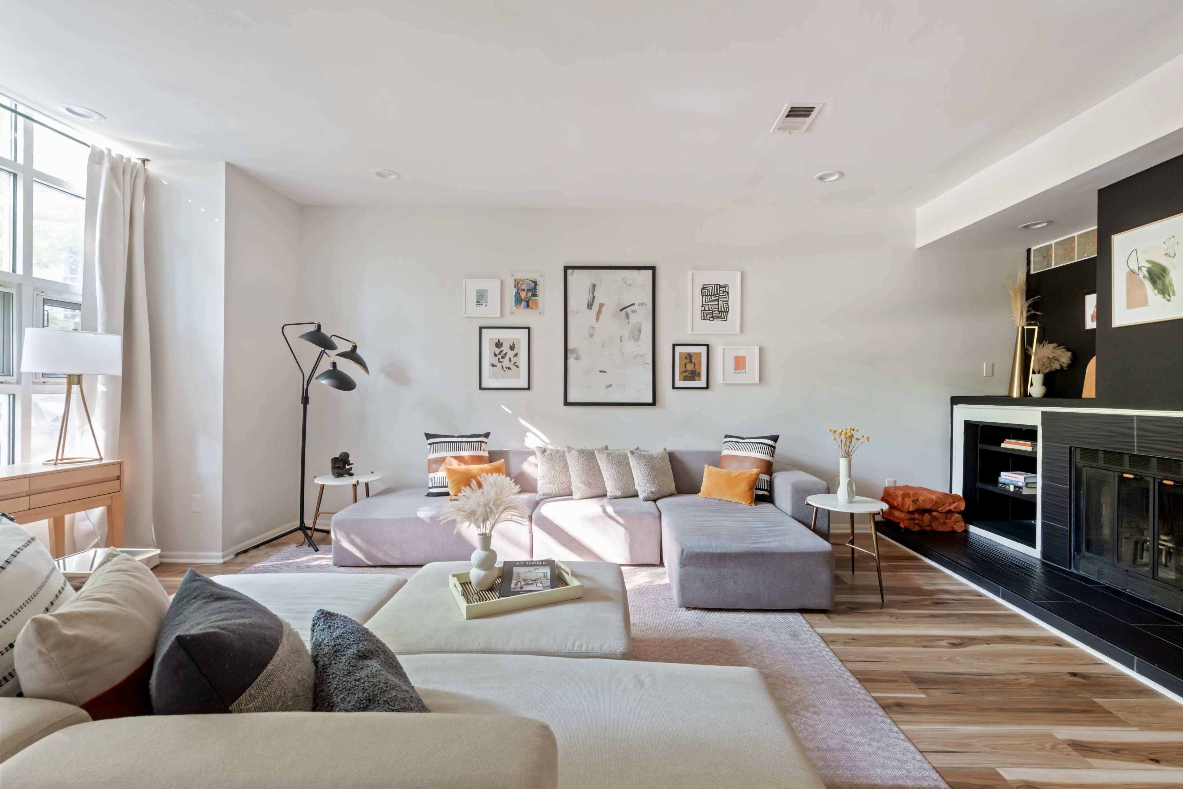 The image shows a modern living room with a light gray color scheme, featuring a large sectional sofa, a coffee table, and various artworks on the walls.