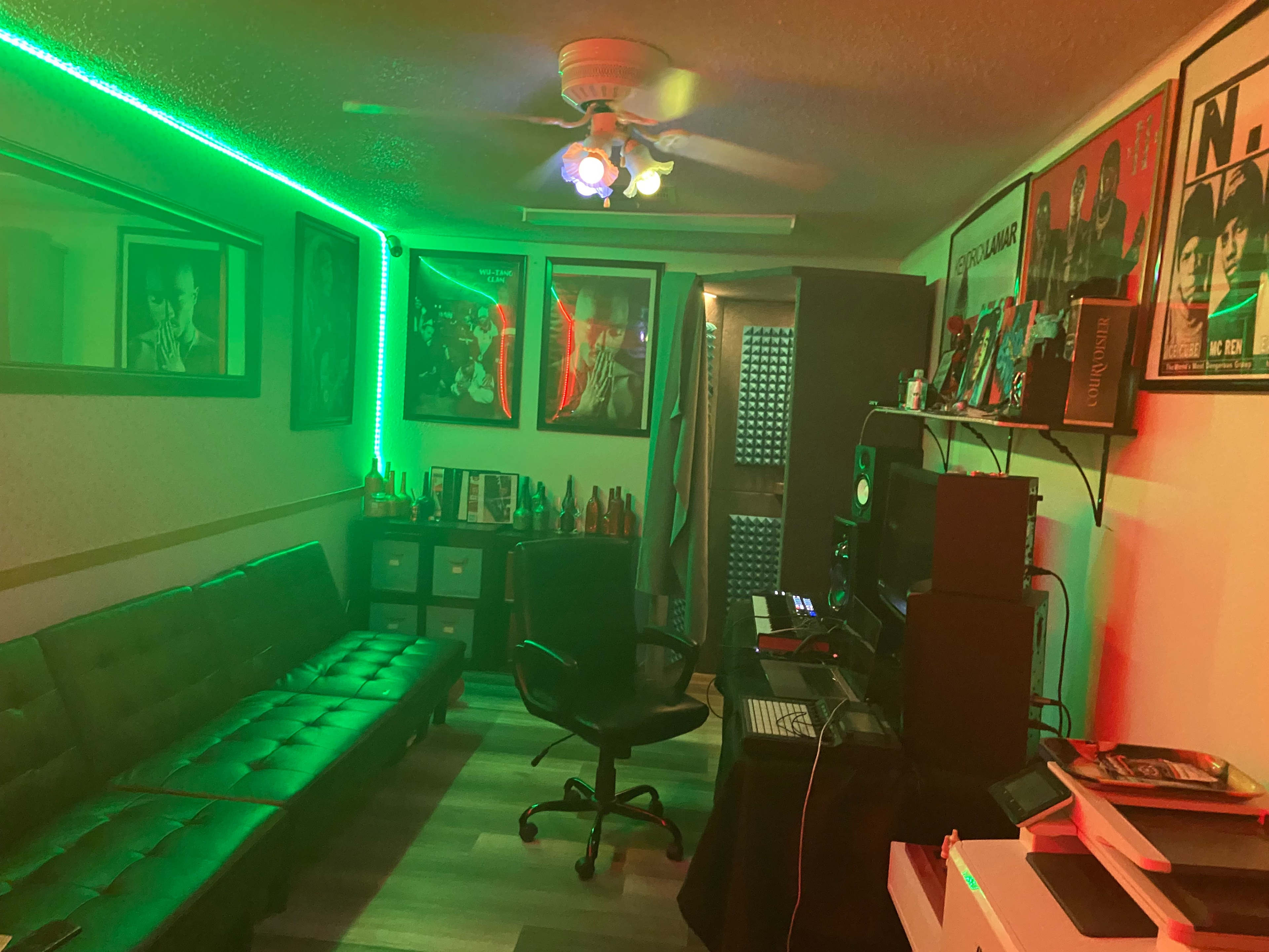 A dimly lit room with green LED lights, featuring a black sofa, a desk with audio equipment, and framed posters on the walls.