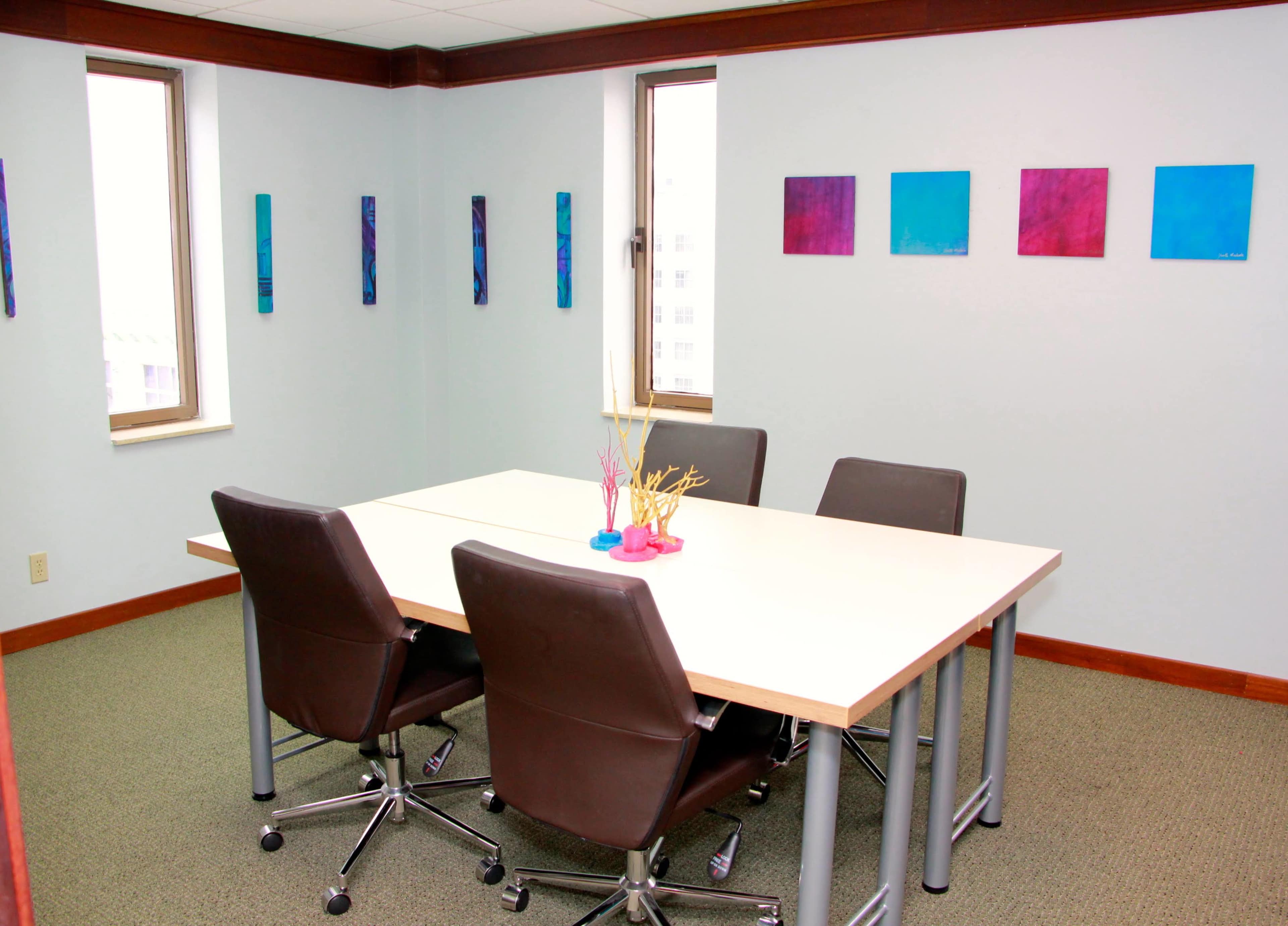 A small conference room features a rectangular table surrounded by four chairs, with colorful paintings on the walls.
