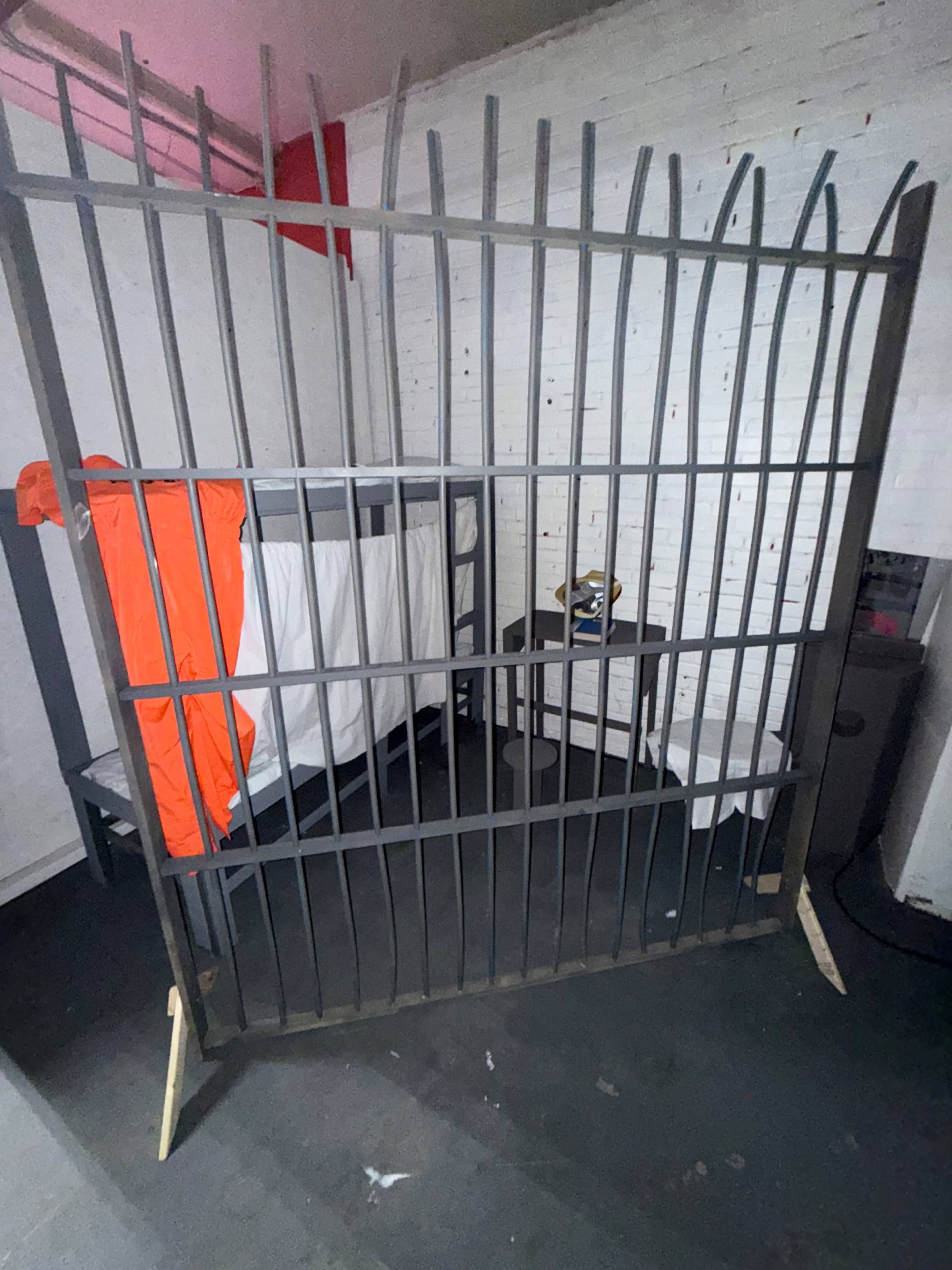 The image shows a prison cell with metal bars, a bunk bed, a chair, and an orange jumpsuit hanging on the wall.