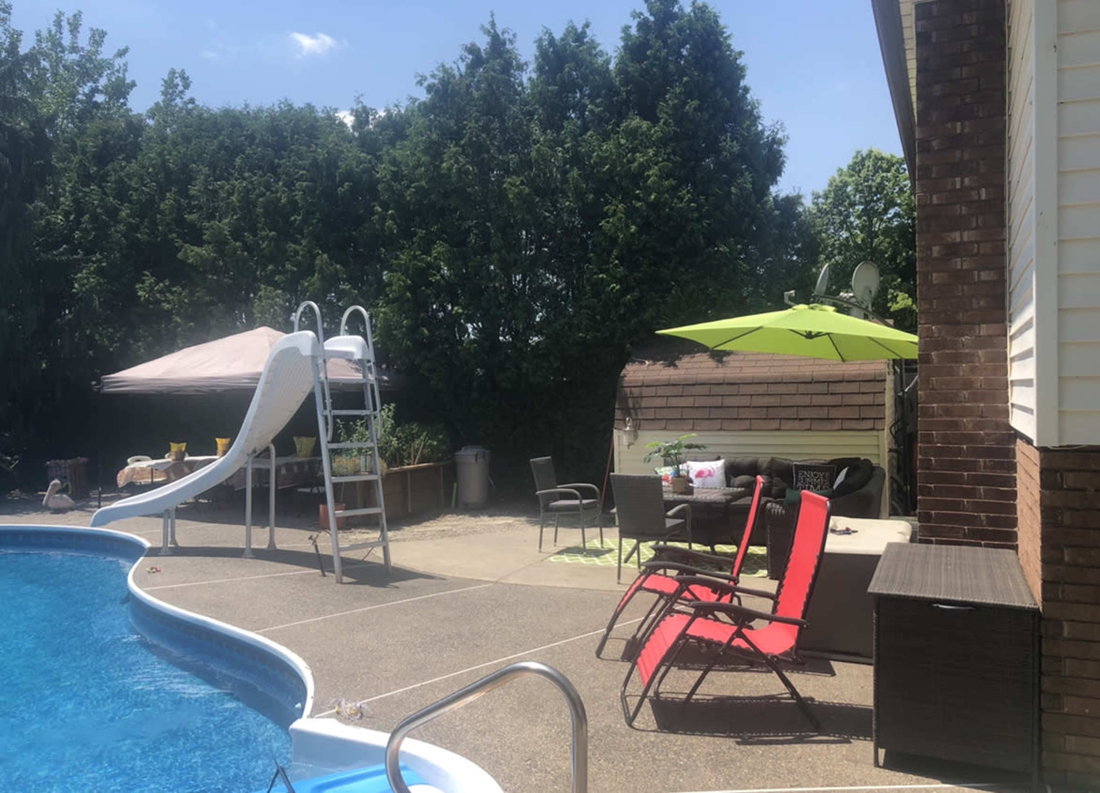 A backyard pool area featuring a slide, a green umbrella, lounge chairs, and a covered seating area surrounded by trees.