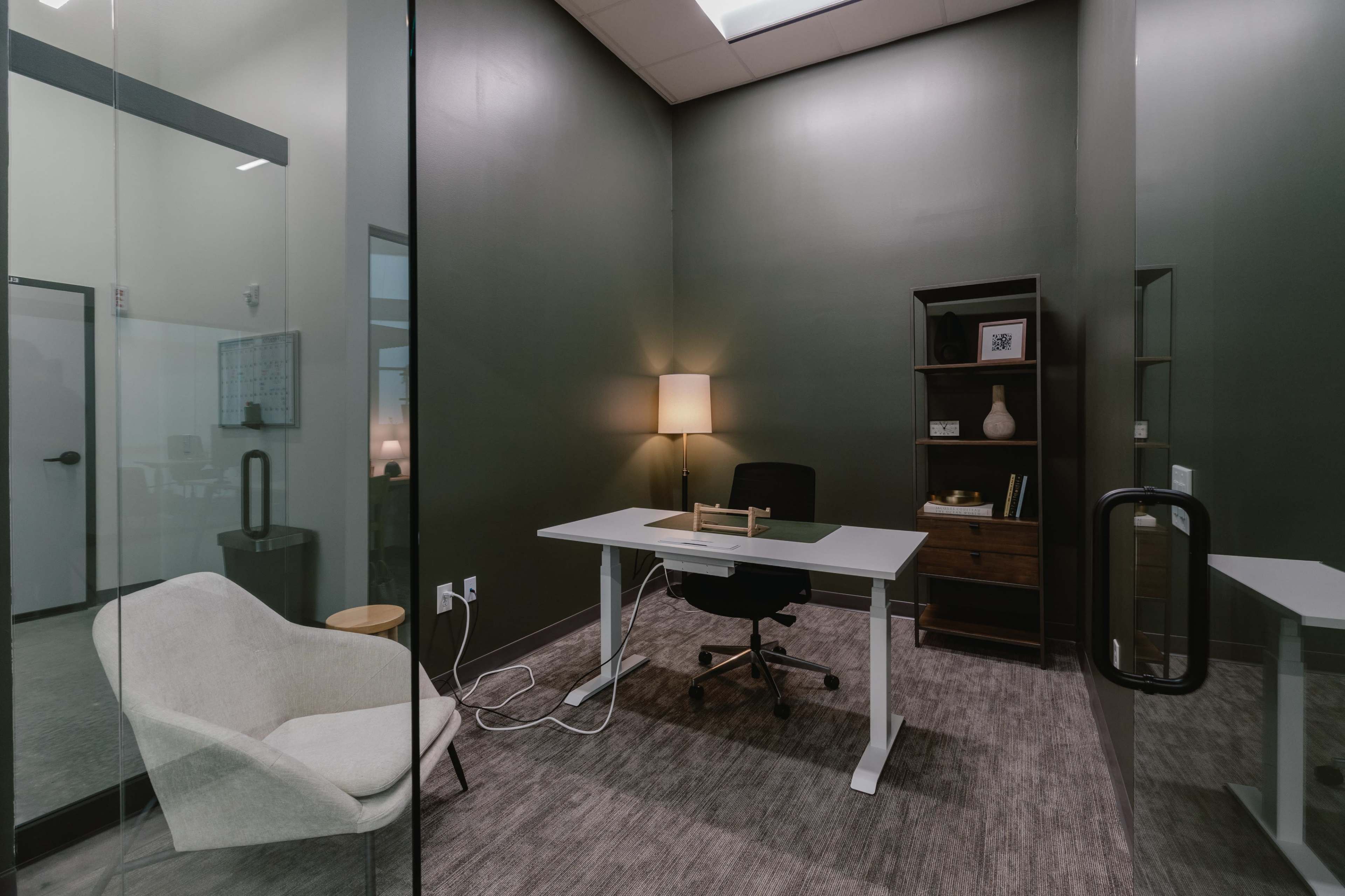 The image shows a small office space with a desk, an office chair, a lamp, and a bookshelf against a dark green wall.