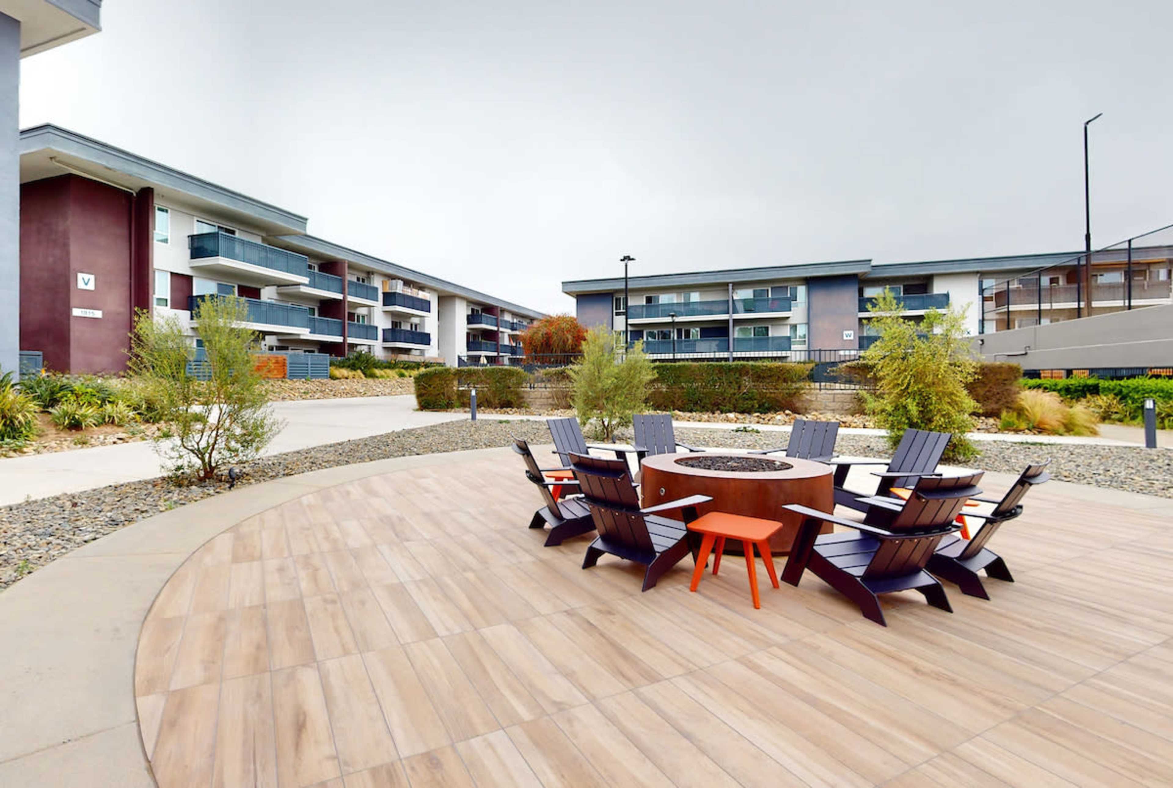 A circular fire pit surrounded by wooden chairs is situated in a landscaped courtyard with modern apartment buildings in the background.