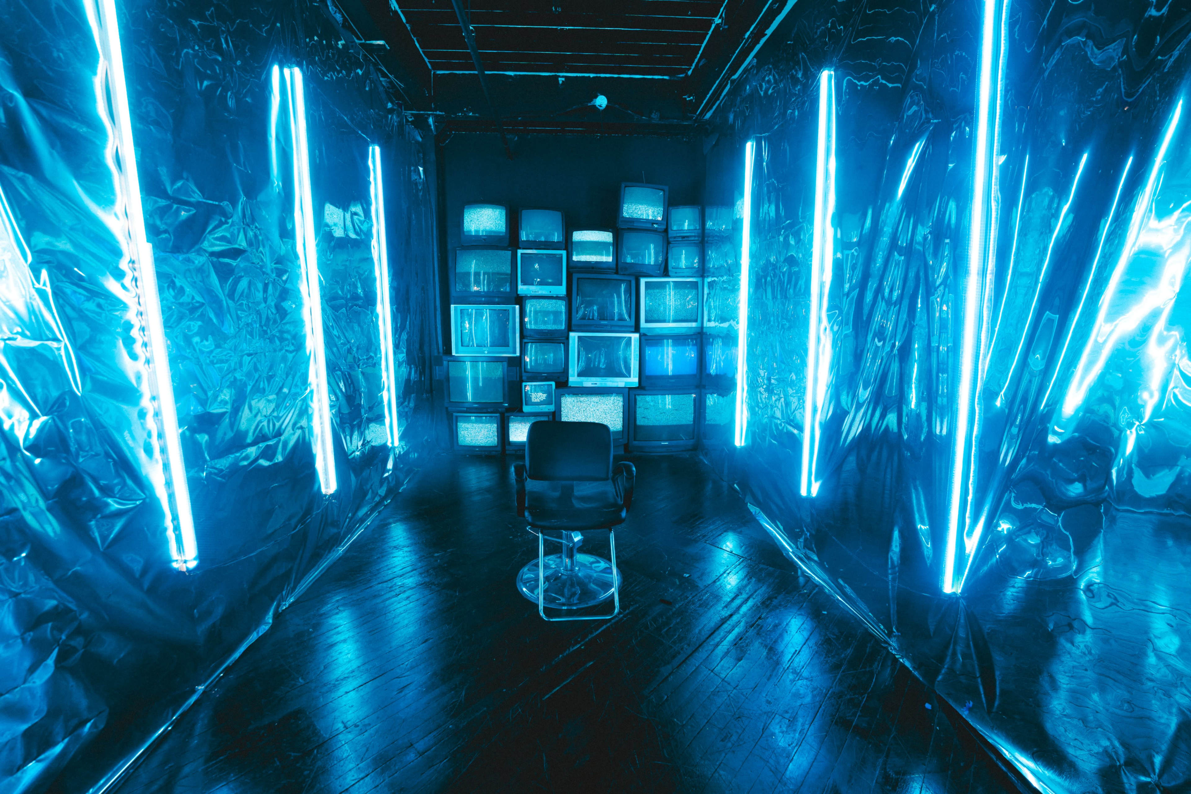 The scene features a dimly lit room with reflective plastic walls, illuminated by blue neon lights and filled with vintage televisions stacked in the background.
