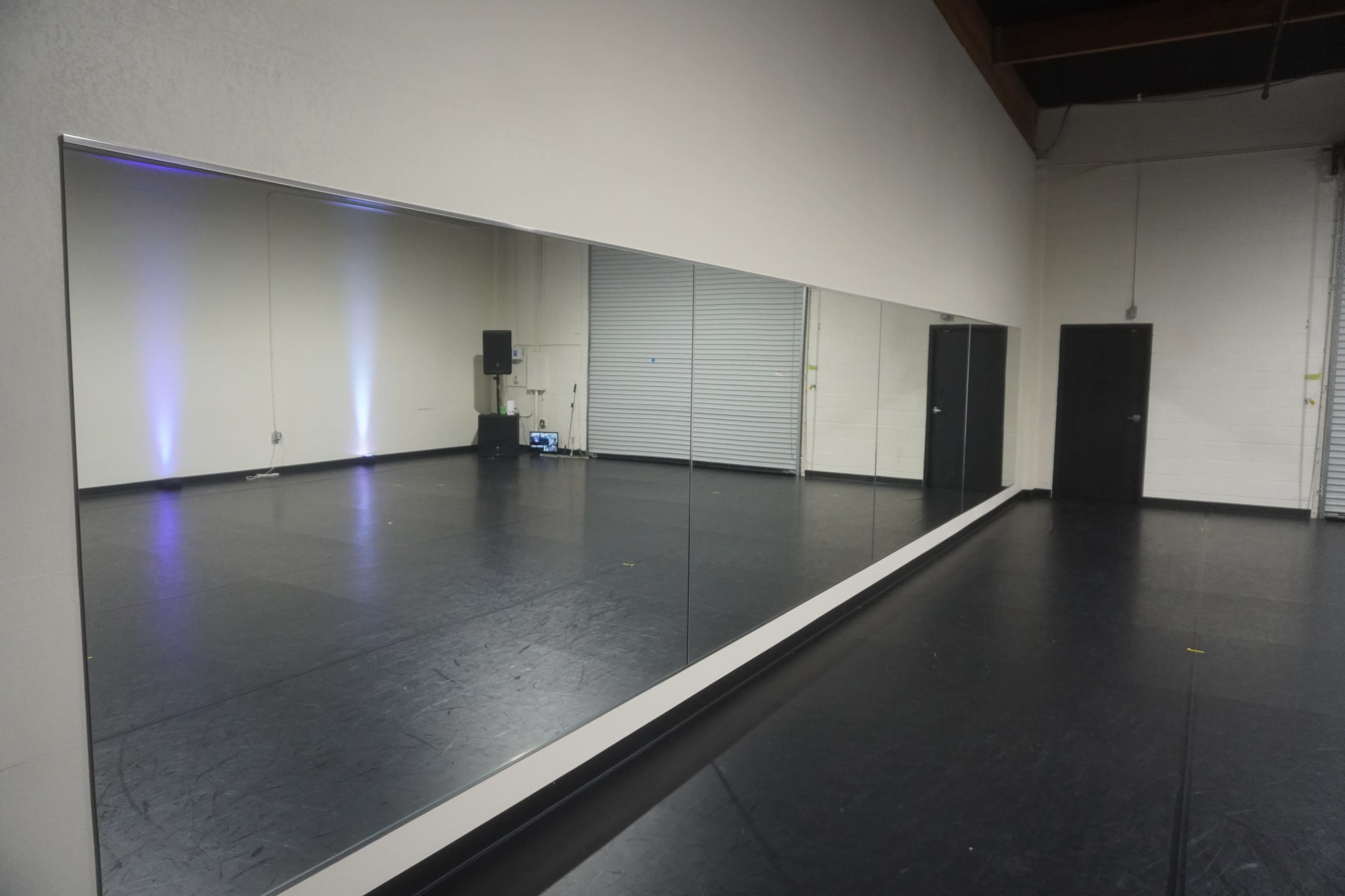 A large wall mirror reflecting a spacious, empty studio with a black floor and a closed door at one end.