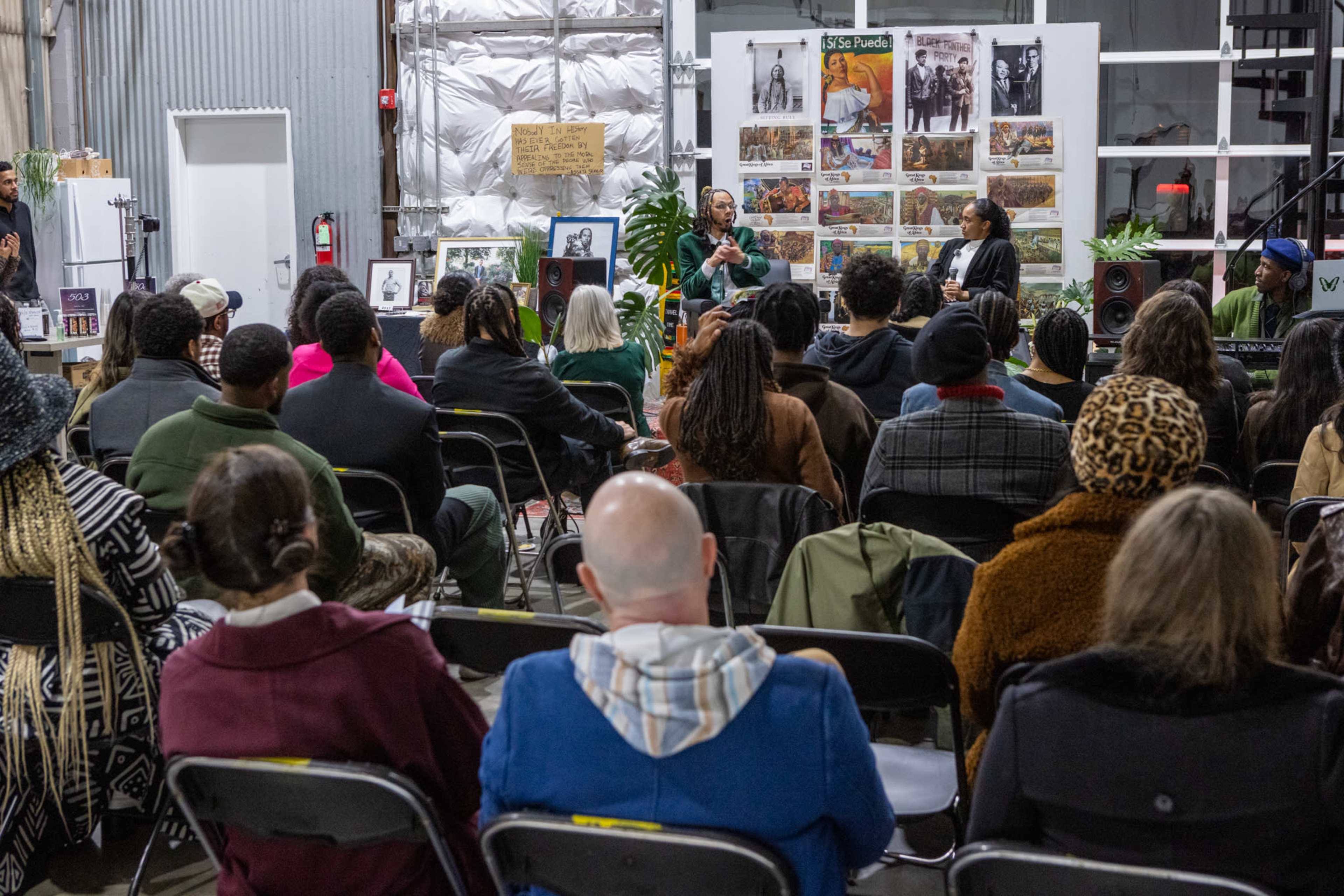 A diverse group of people sits in a warehouse, engaged in a discussion at the front, surrounded by photographs and art on the walls.