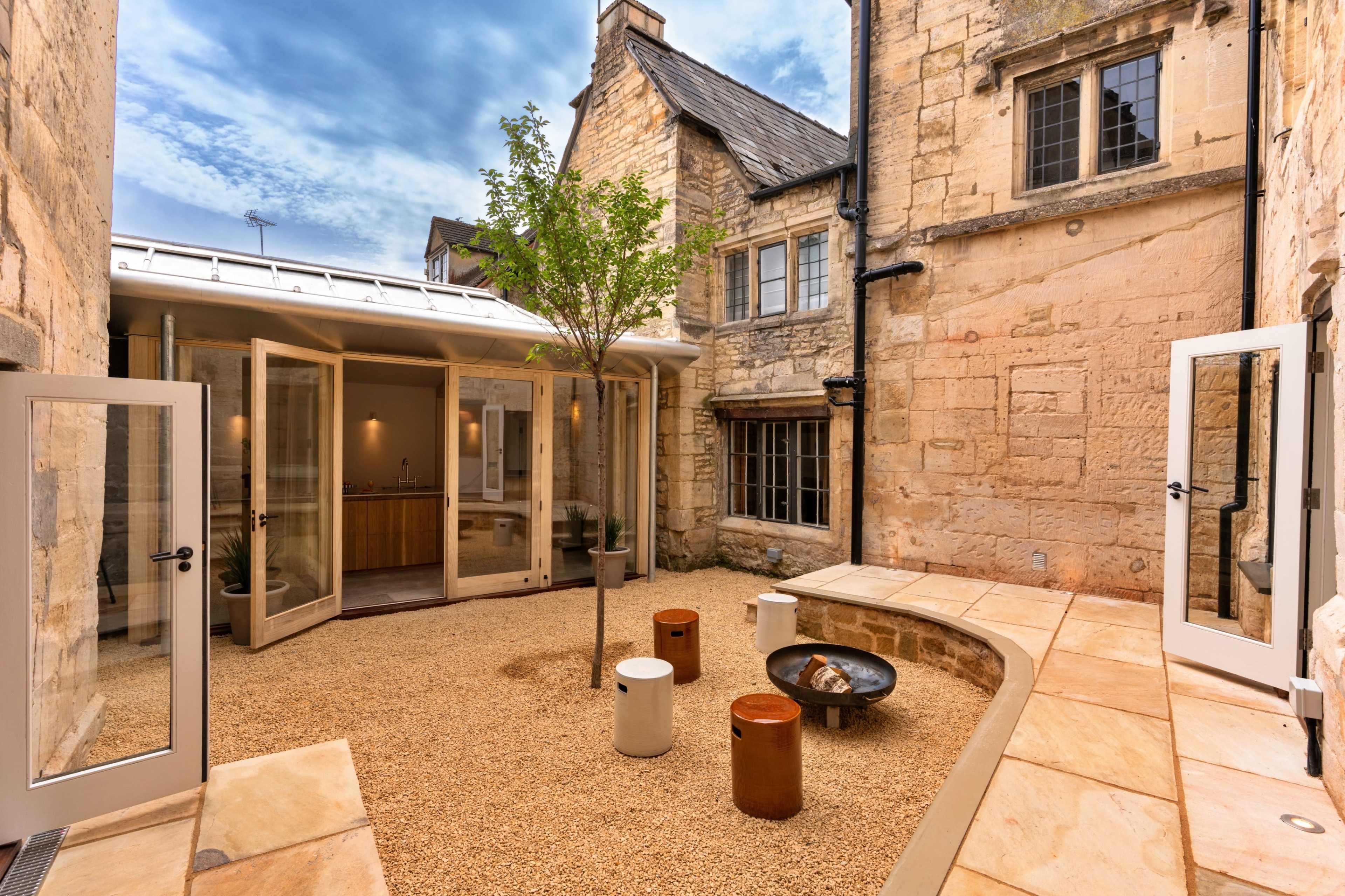 A stone courtyard surrounded by buildings, featuring a central tree, seating elements, and a fire pit.