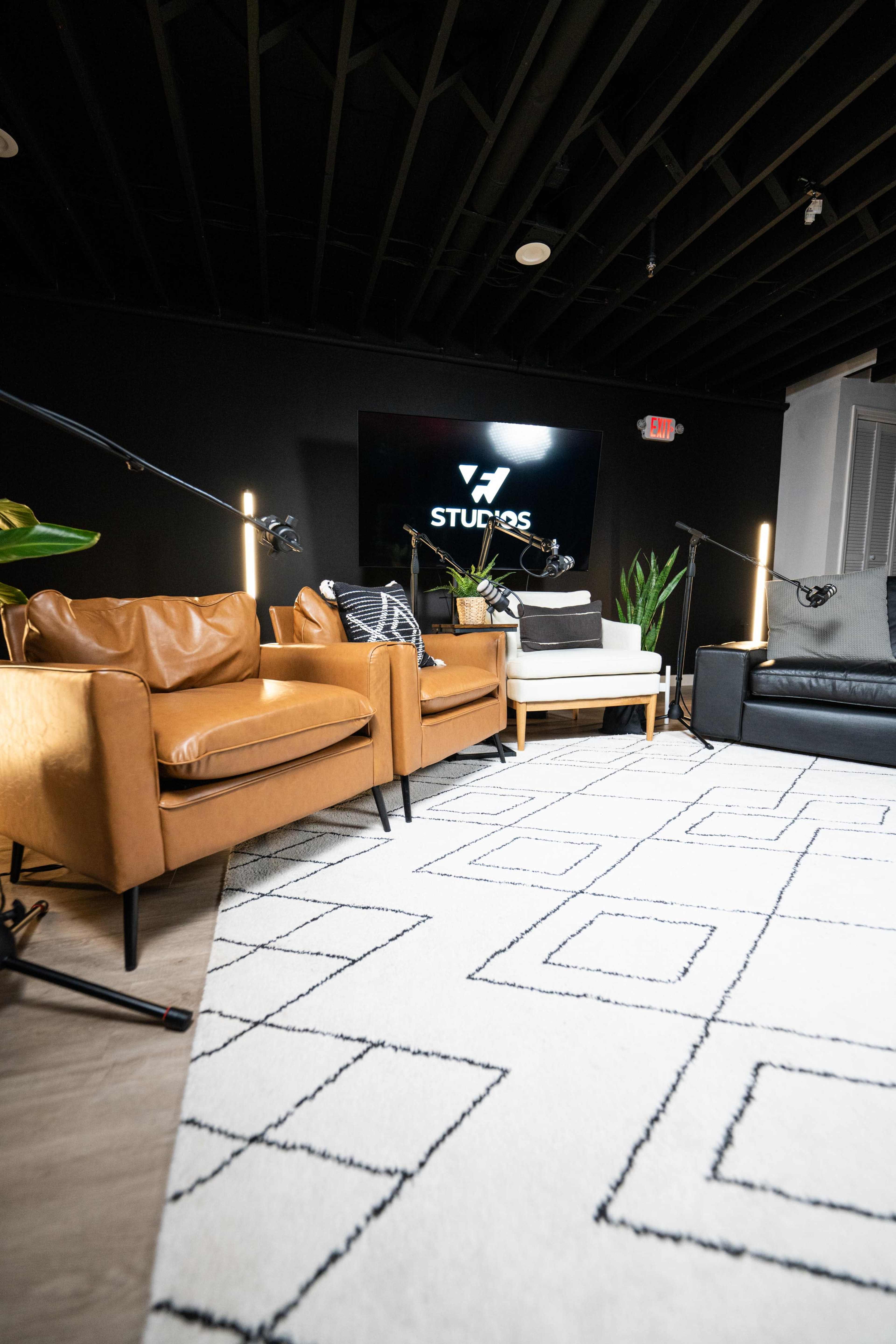 The image shows a modern interior setting with two brown leather chairs, a black couch, a light-colored coffee table, and a patterned rug, all arranged under a black ceiling with studio lighting.