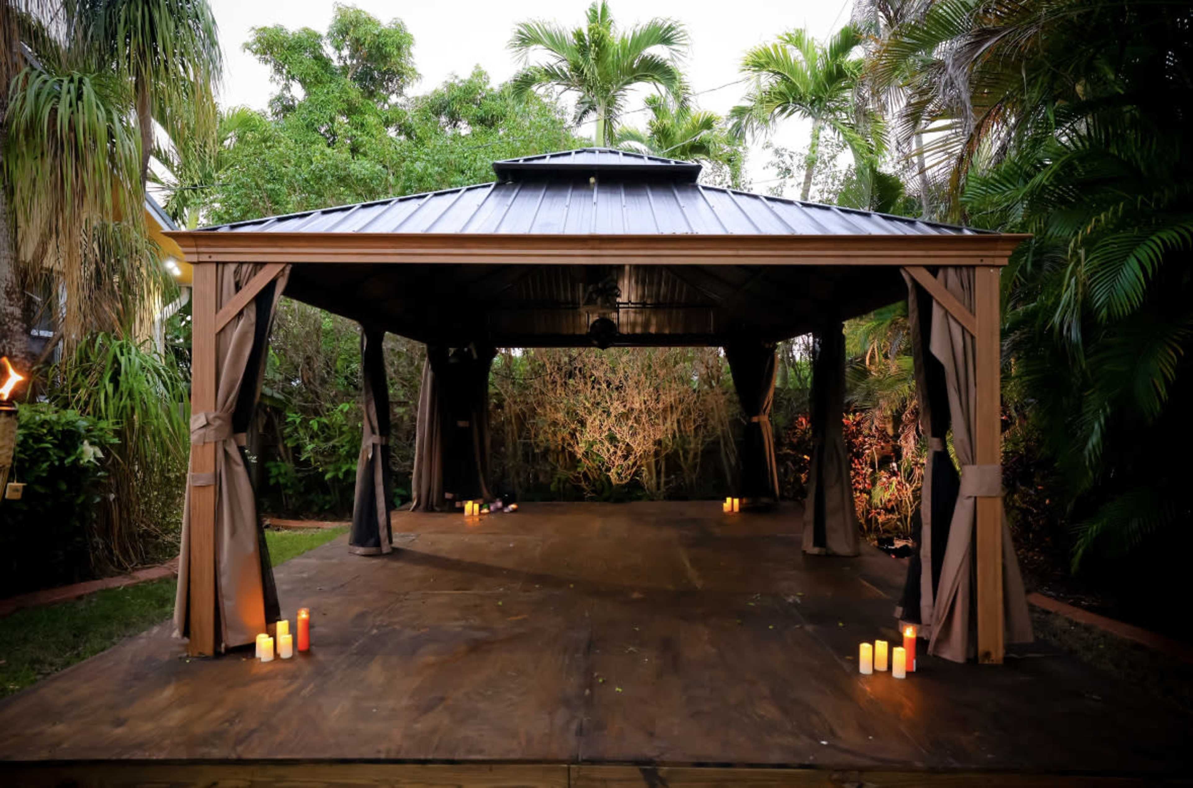 An outdoor pavilion with a dark wooden floor is surrounded by lush greenery and illuminated by candles placed on the ground.