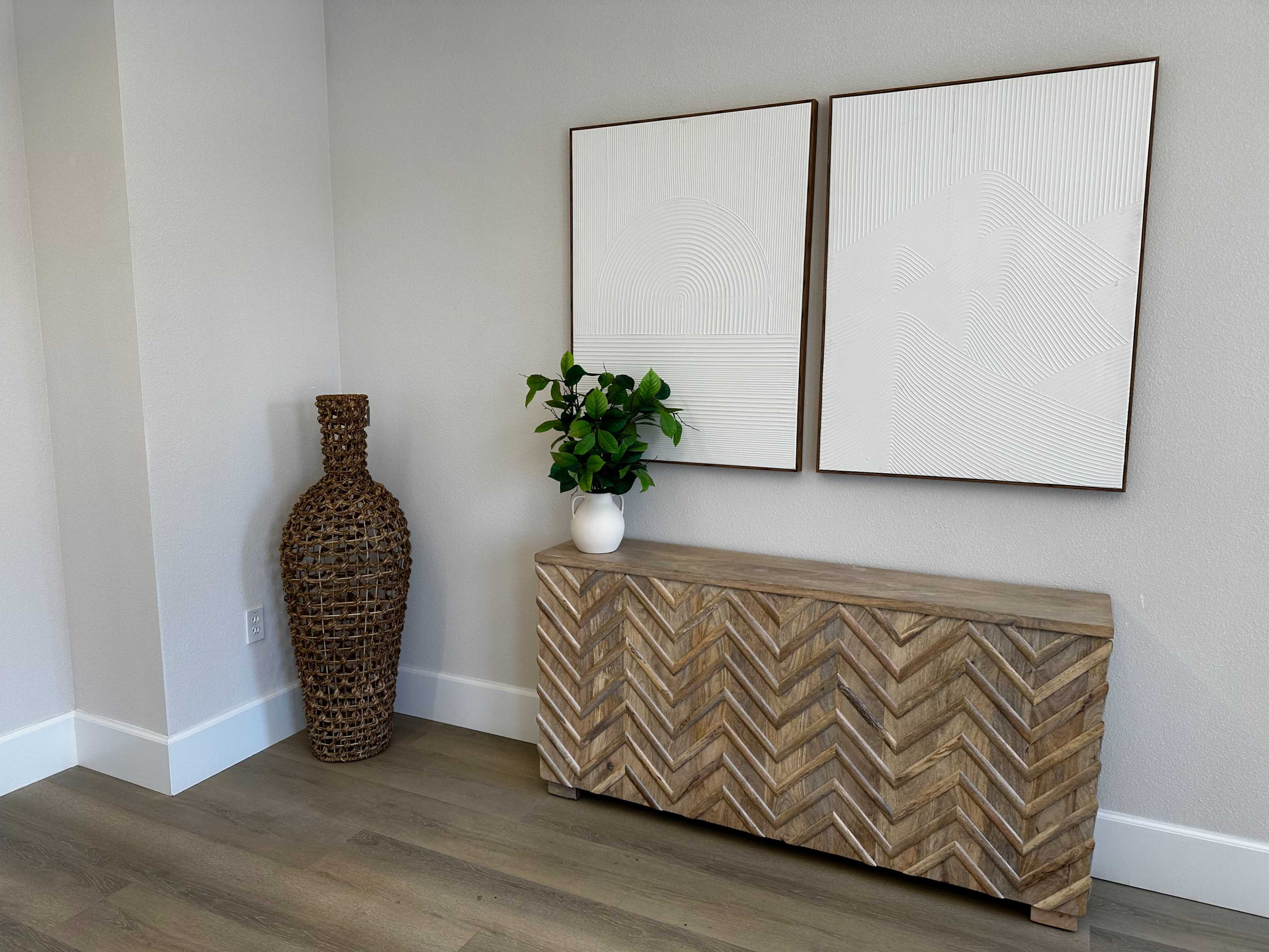 The scene features a wooden console table with a chevron pattern, accompanied by two framed abstract artworks and a woven vase with a plant beside it.