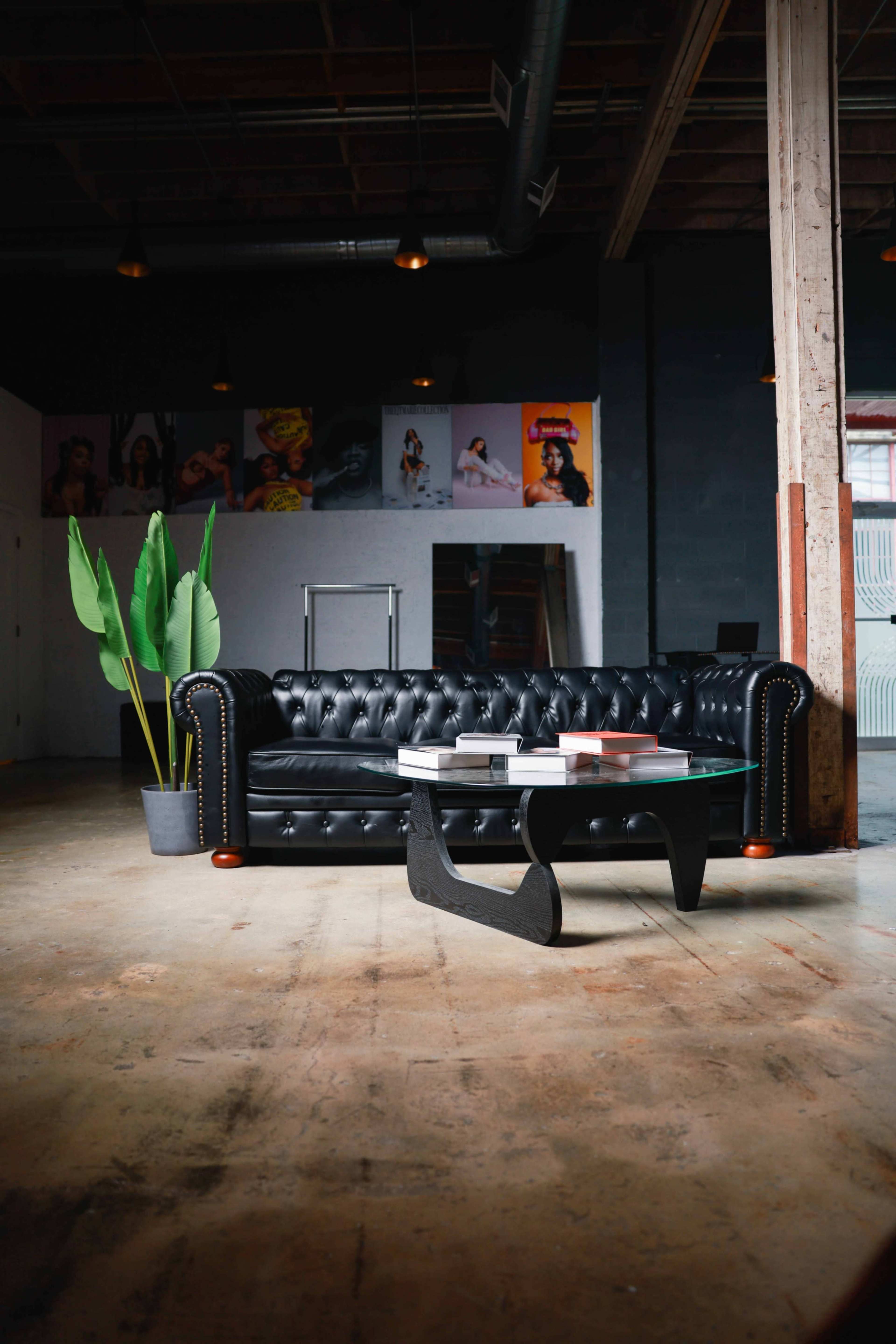 The image features a black tufted leather couch paired with a modern coffee table, situated in an industrial-style room with exposed wooden beams and colorful art on the walls.