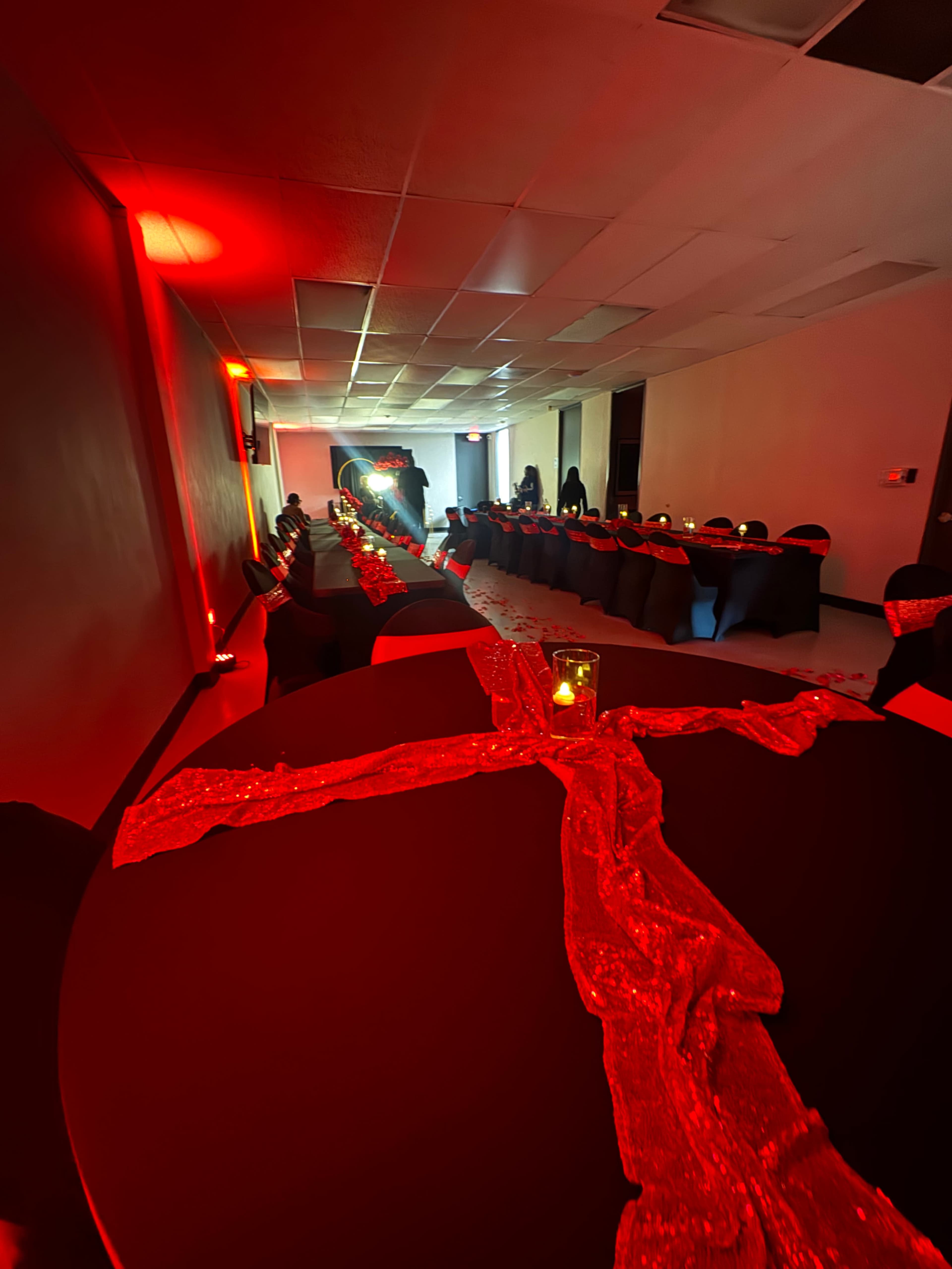 A long table covered with black cloth and adorned with a red runner is set up in a dimly lit room, with candles illuminating the scene.