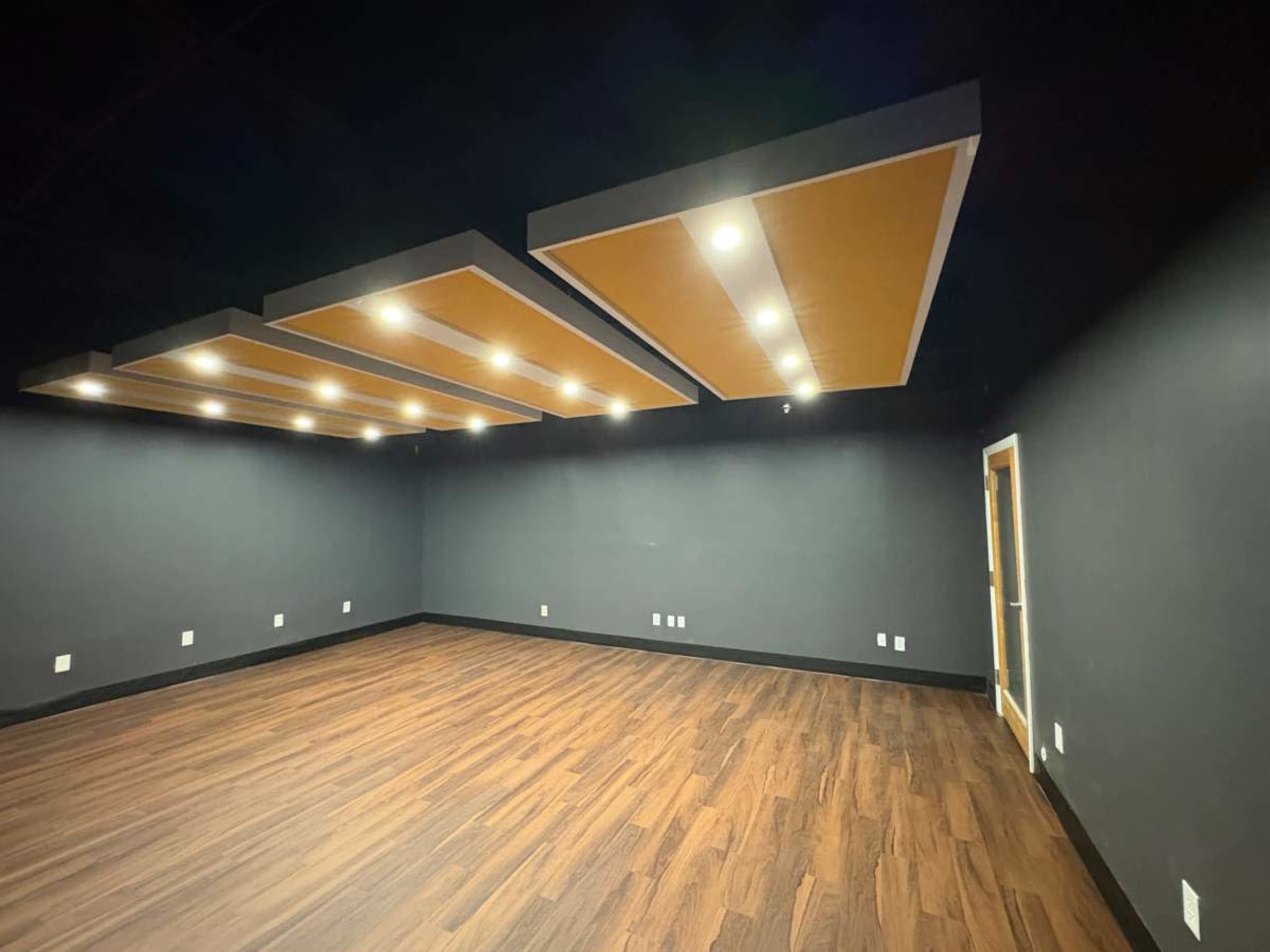A spacious room with dark gray walls, wooden flooring, and three large, illuminated ceiling panels.