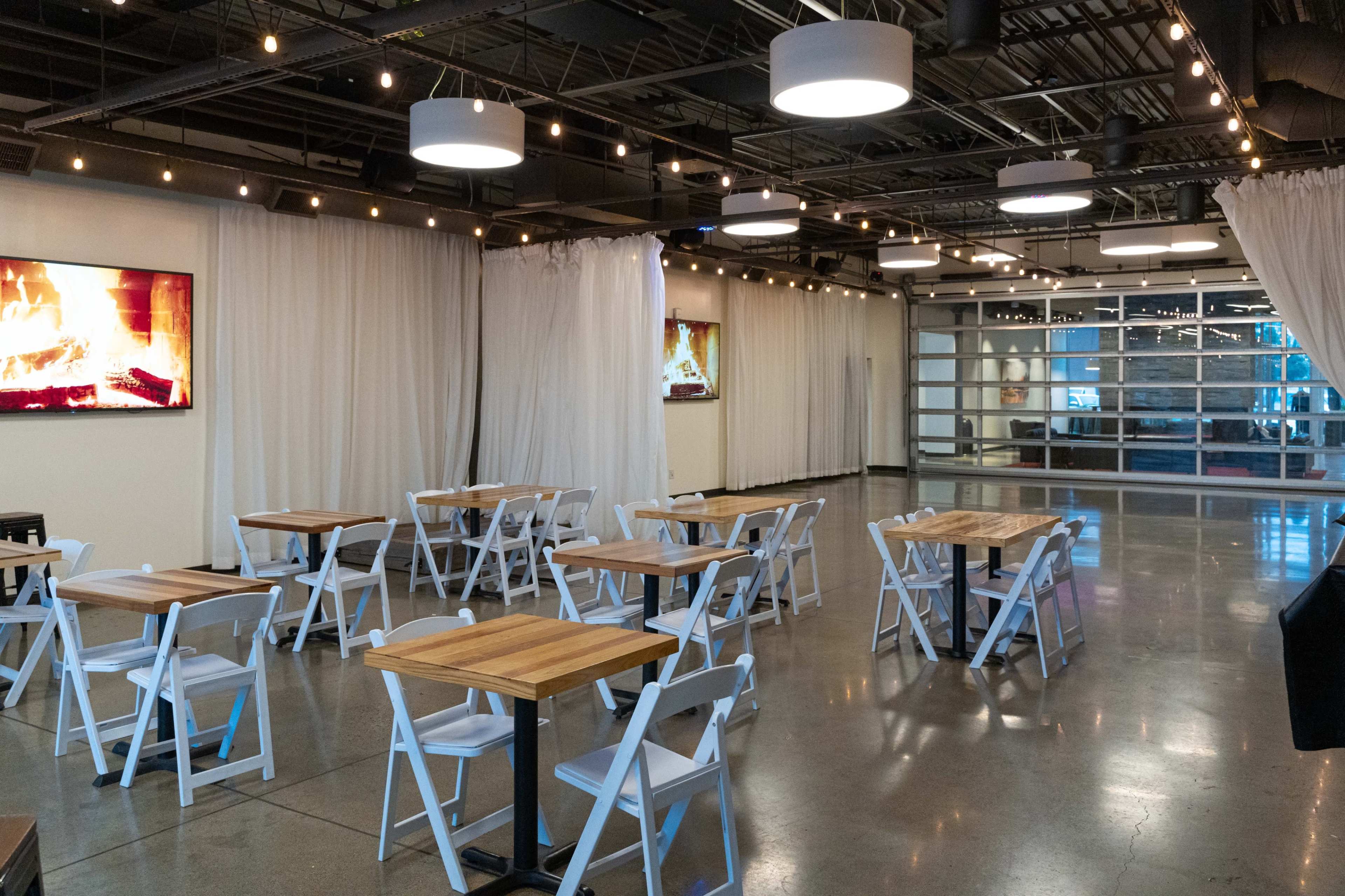 A spacious interior of a modern venue featuring white tables, folding chairs, and ambient lighting, with a large screen displaying a fireplace.