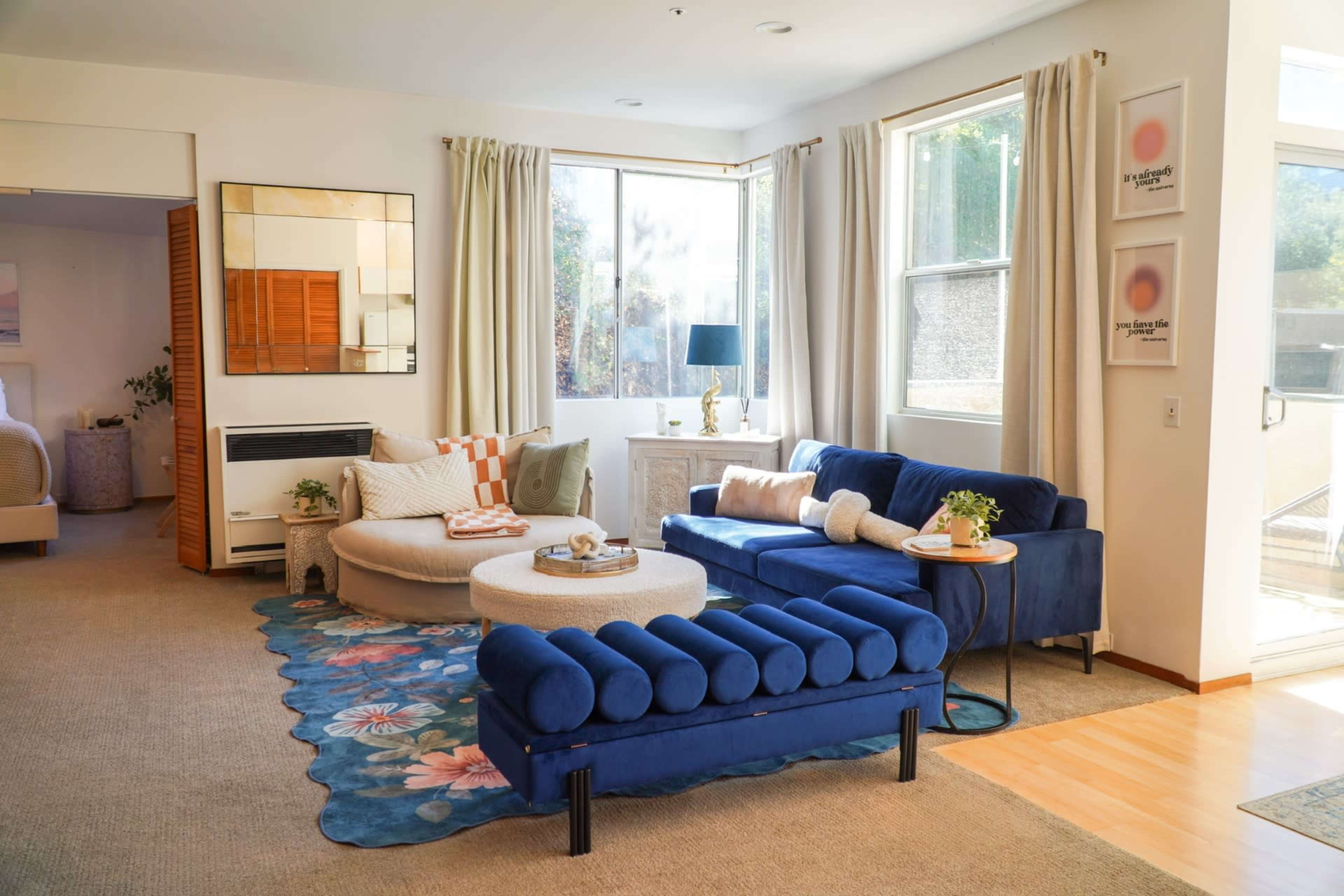 The living room features a blue sectional sofa, a round coffee table, and large windows that allow natural light to enter the space.