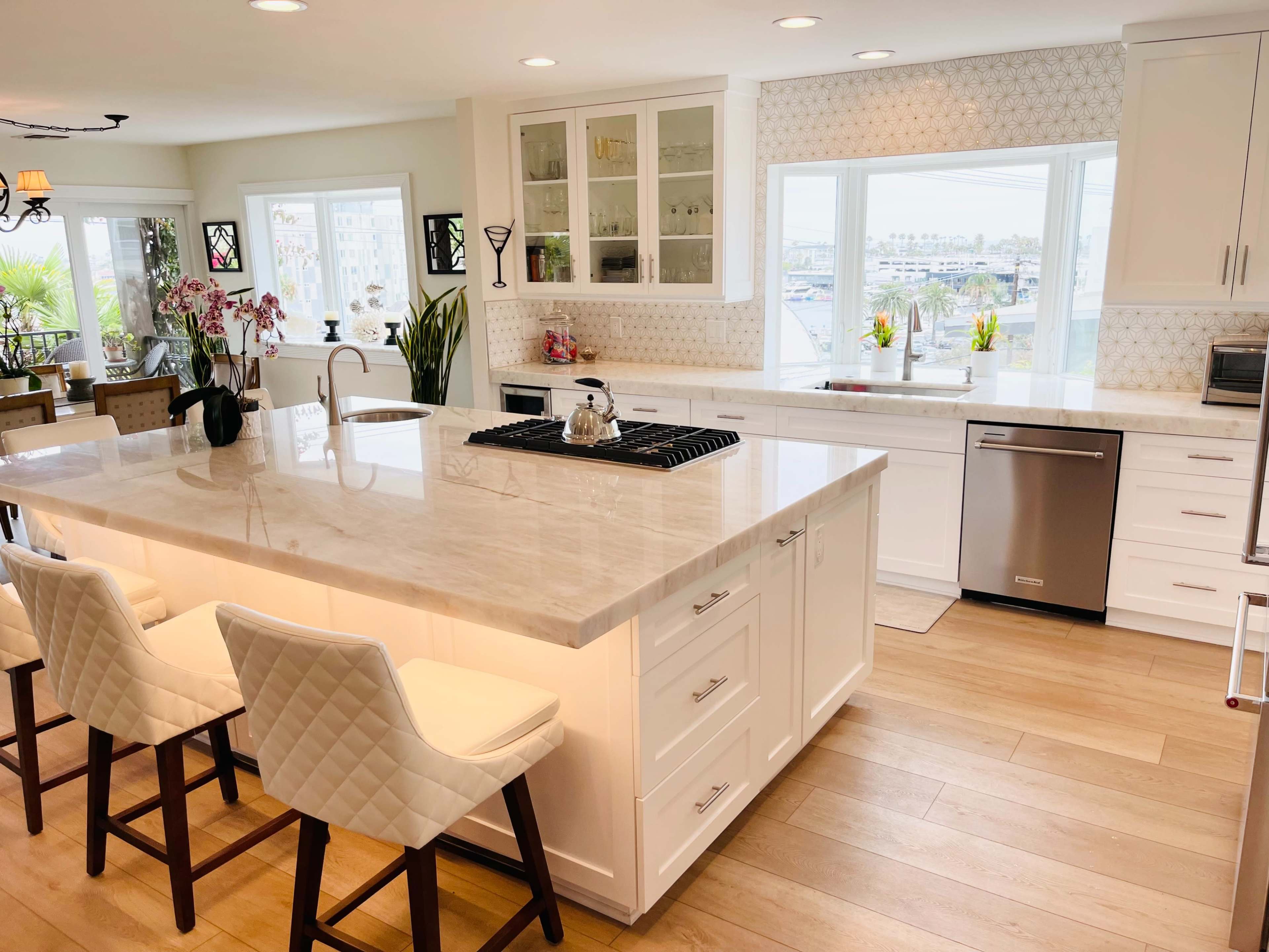 A modern kitchen featuring a large island with a marble countertop, white cabinetry, and a view of an outdoor area through large windows.