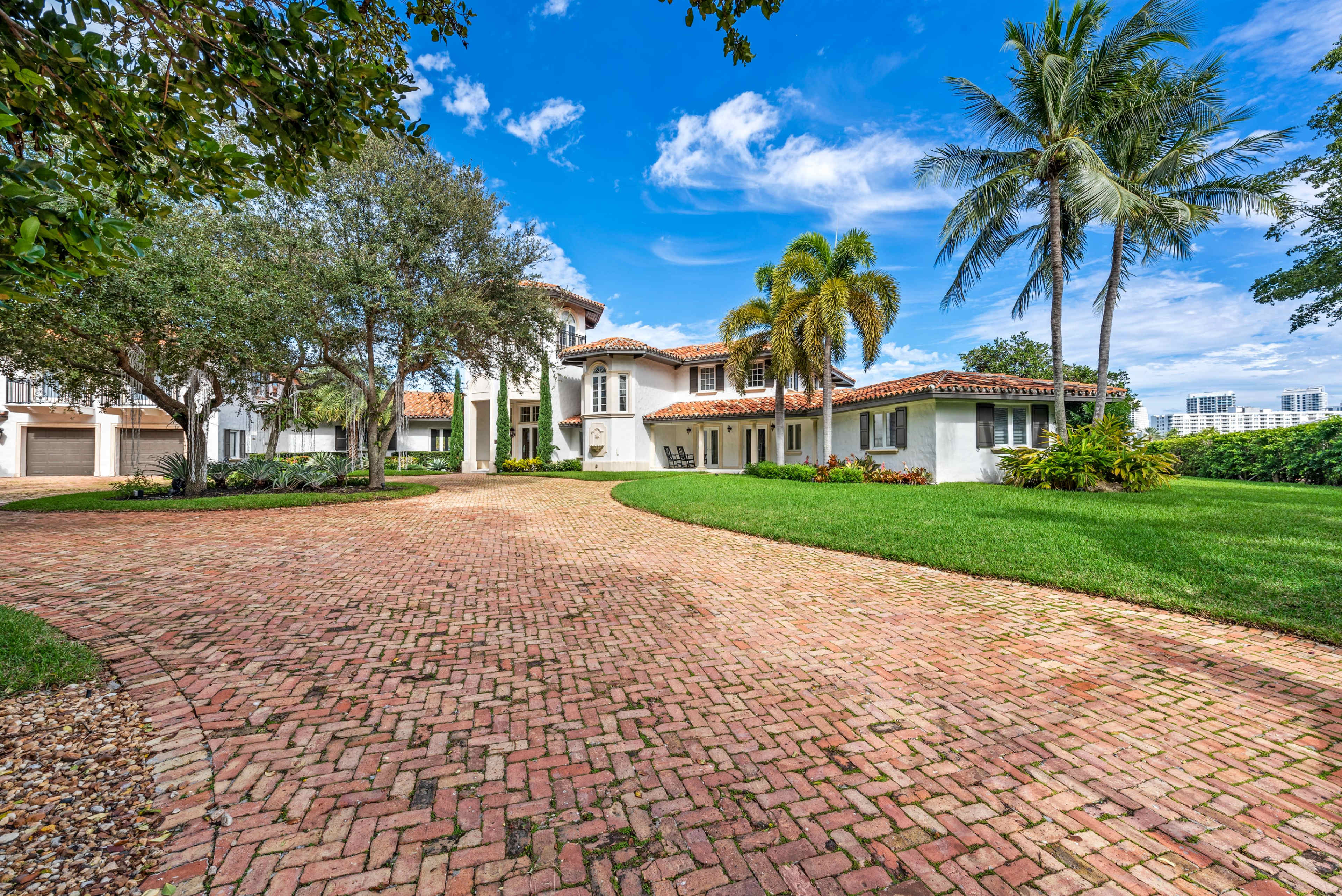 The image shows a large, white stucco house with a tiled roof, surrounded by palm trees and a landscaped yard, and a brick driveway leading to the entrance.