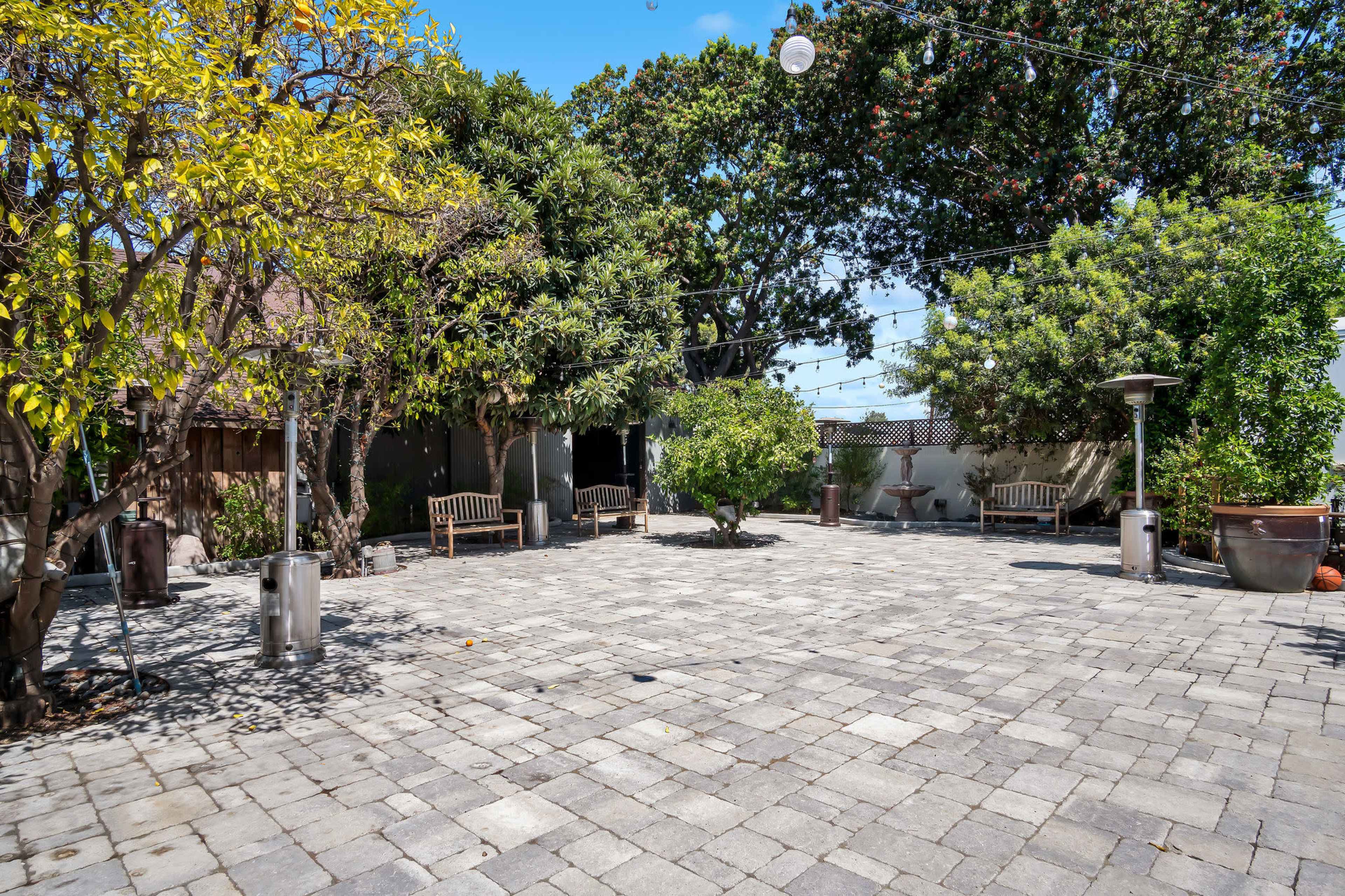The image shows a spacious outdoor courtyard with stone pavers, surrounded by trees and equipped with benches and patio heaters.