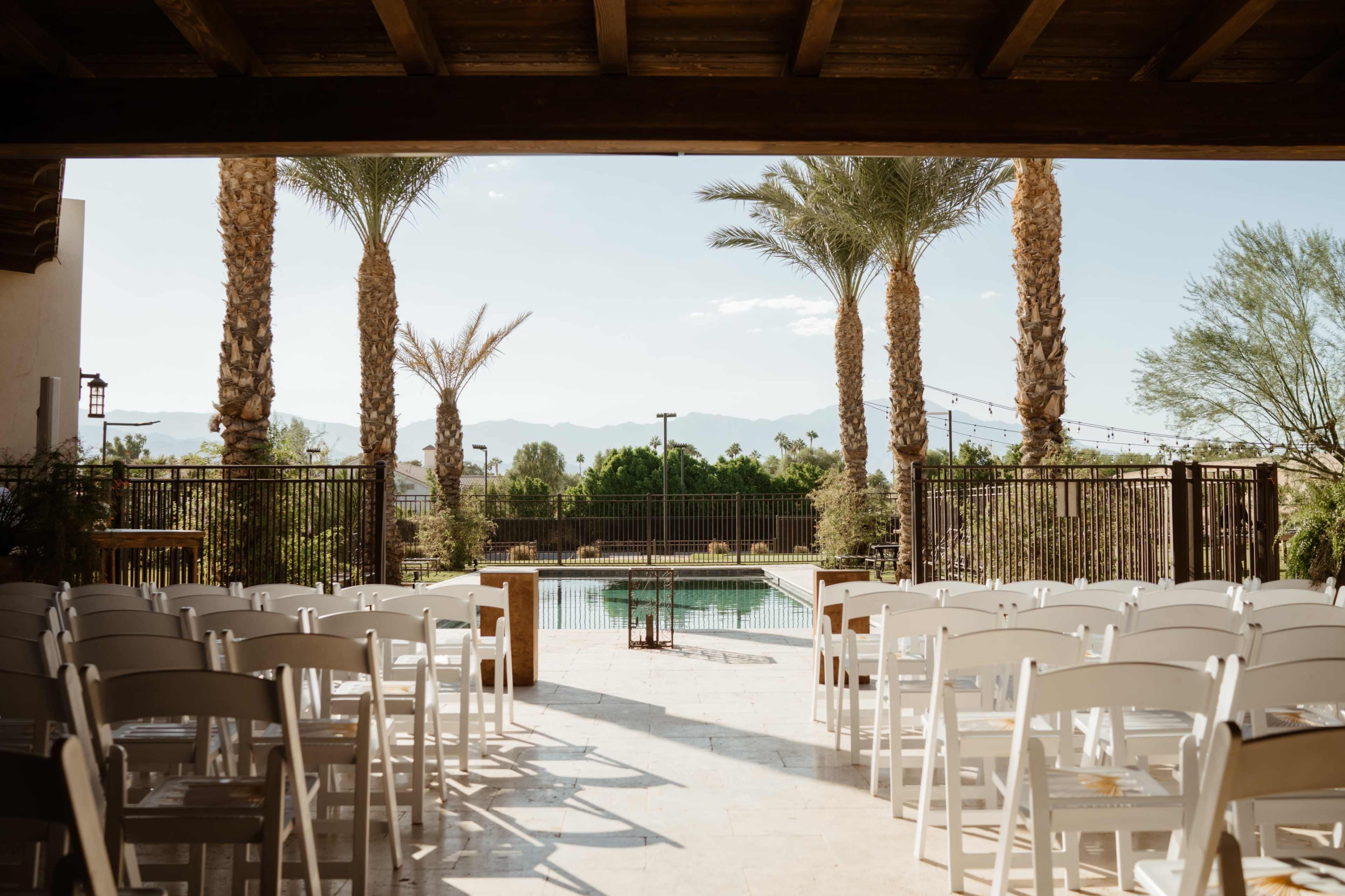 The image shows a view from under a wooden structure towards a swimming pool surrounded by palm trees and mountains in the background, with white chairs arranged for an event.