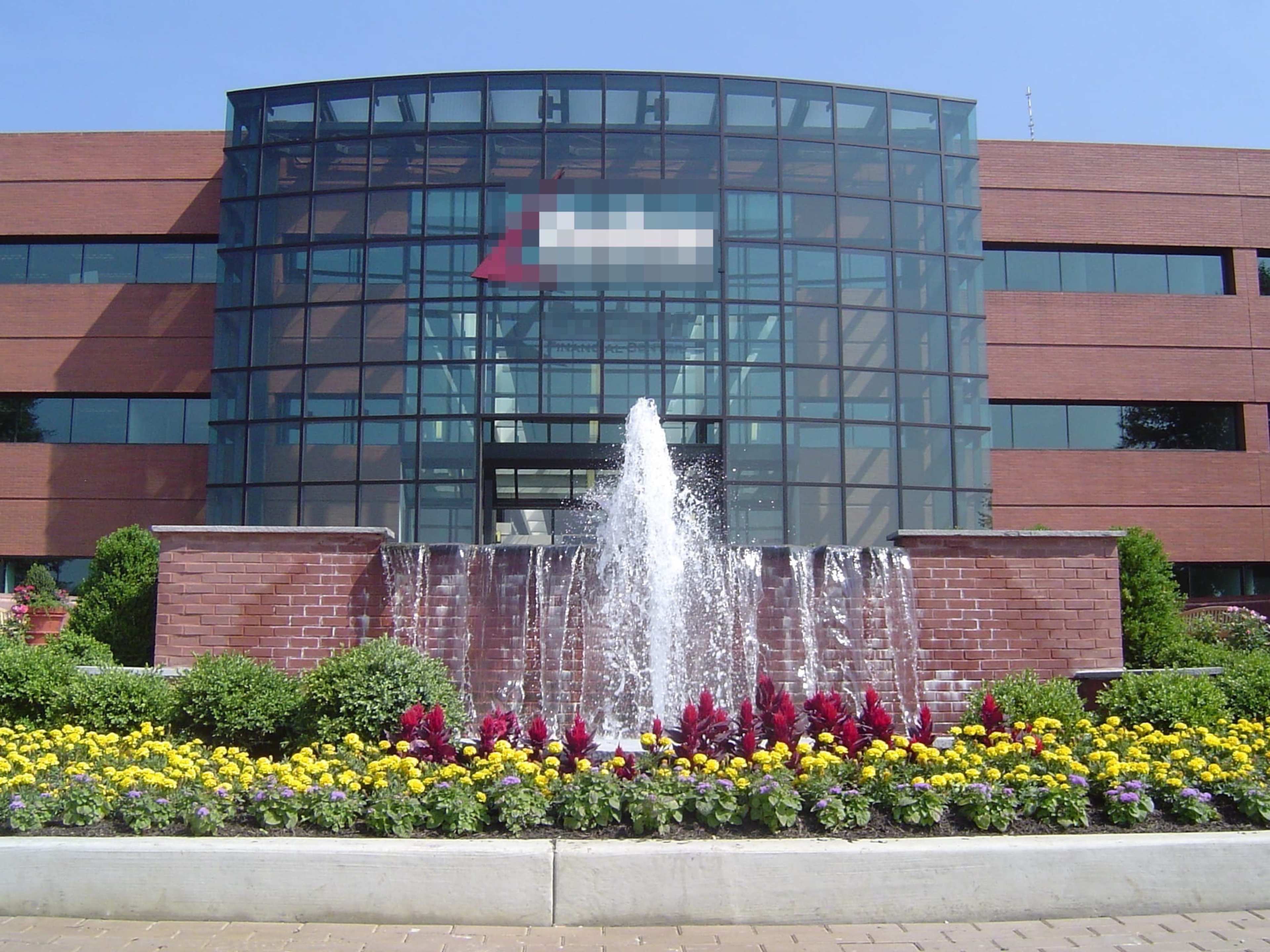A modern brick building with large glass windows, featuring a fountain and a flower bed with colorful plants in the foreground.