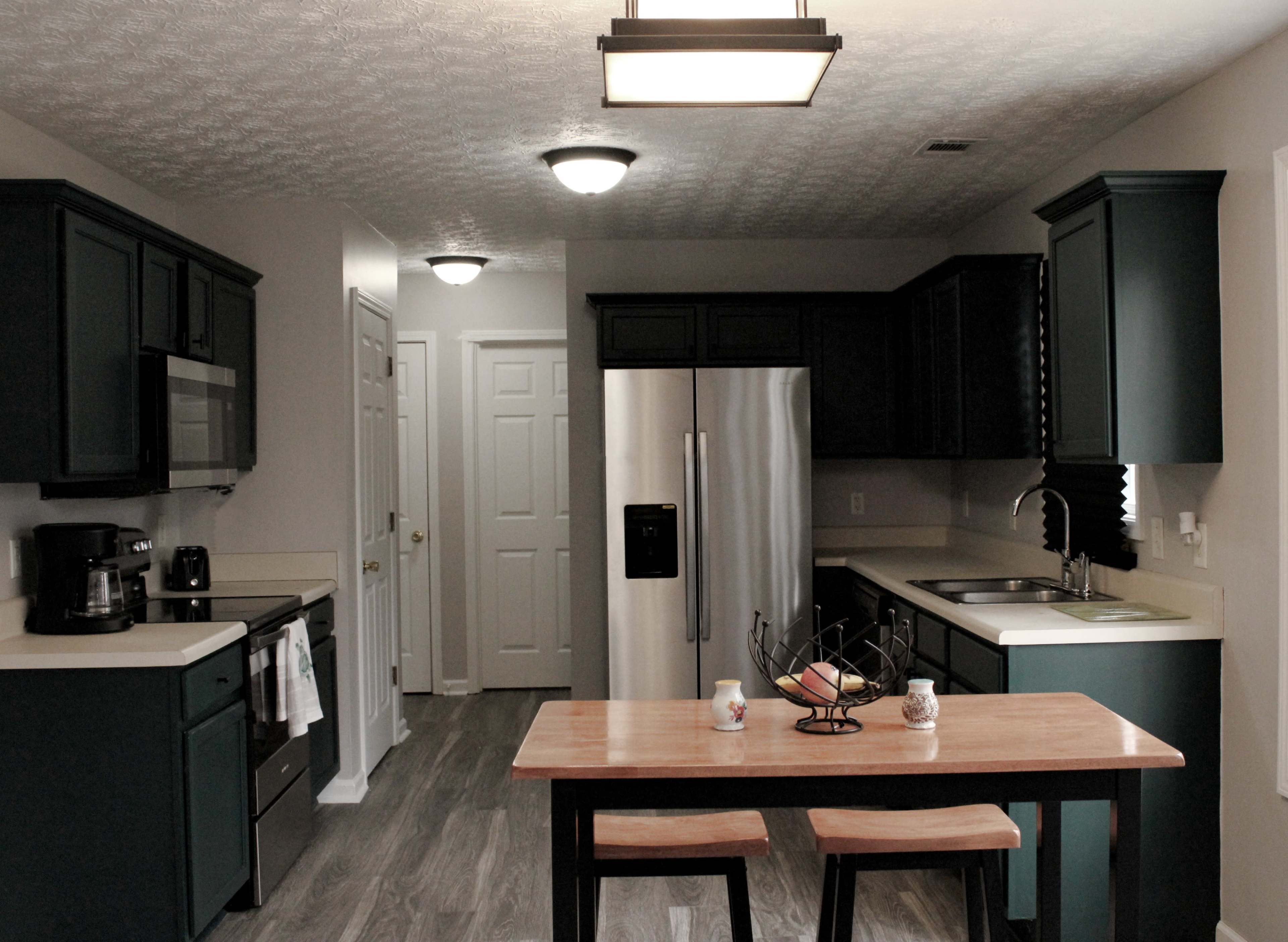 The image shows a modern kitchen featuring dark green cabinets, stainless steel appliances, and a small wooden dining table with a fruit basket.
