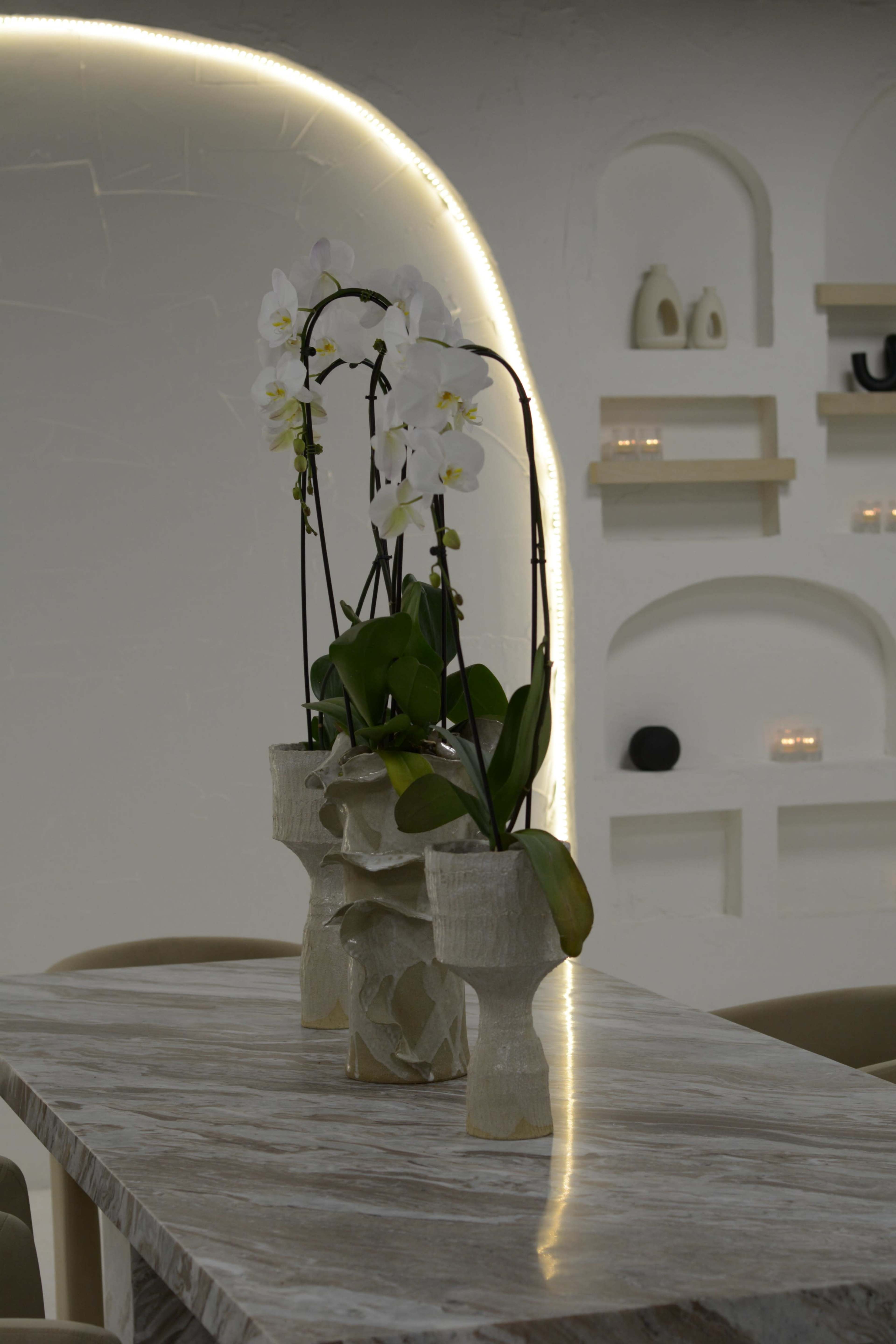 A marble table features two vases holding white orchid flowers, set against a minimalist white wall with shelf decor.