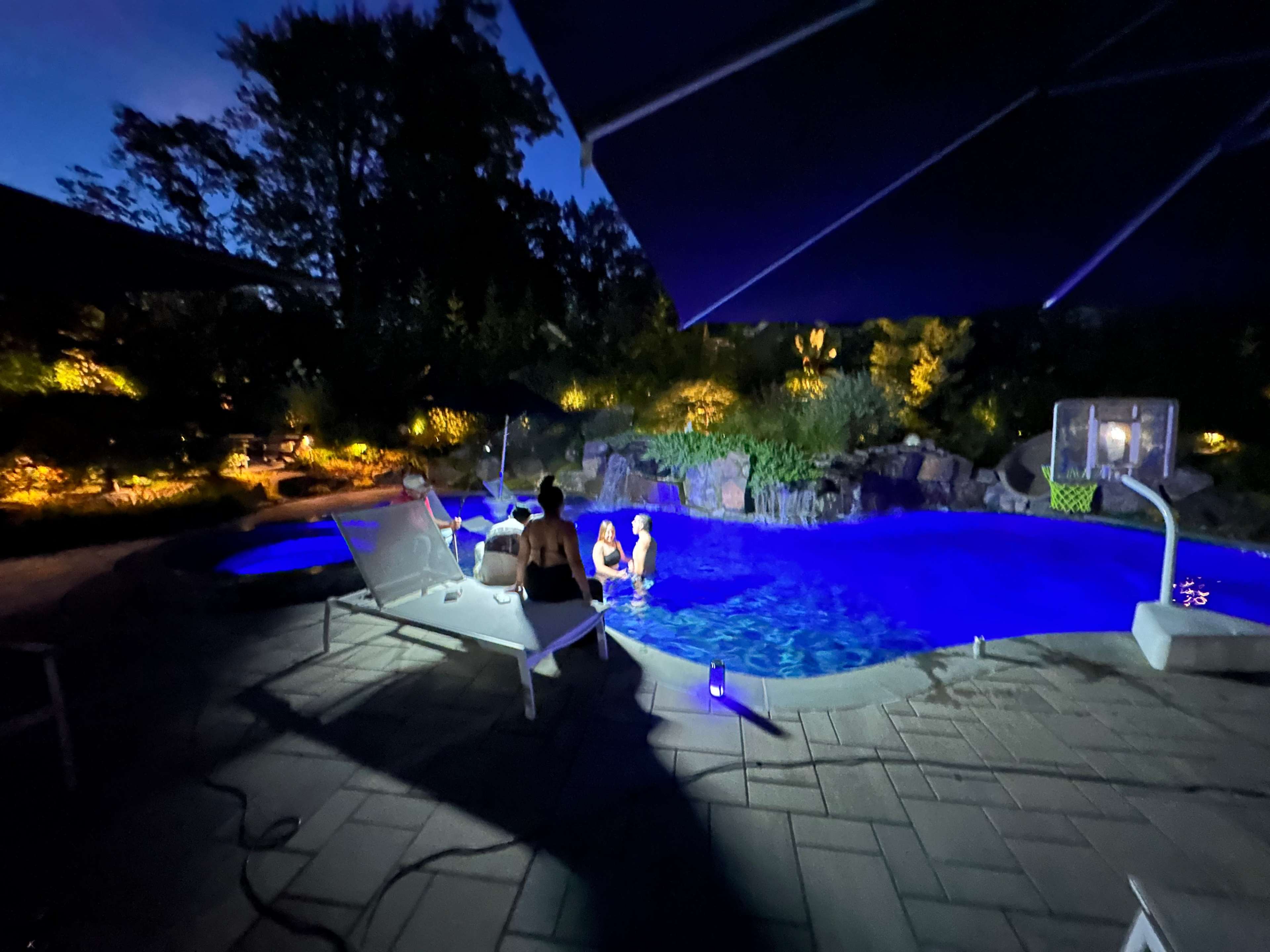 A group of people gathers around a brightly lit pool at night, with surrounding trees and landscaping illuminated by soft lighting.