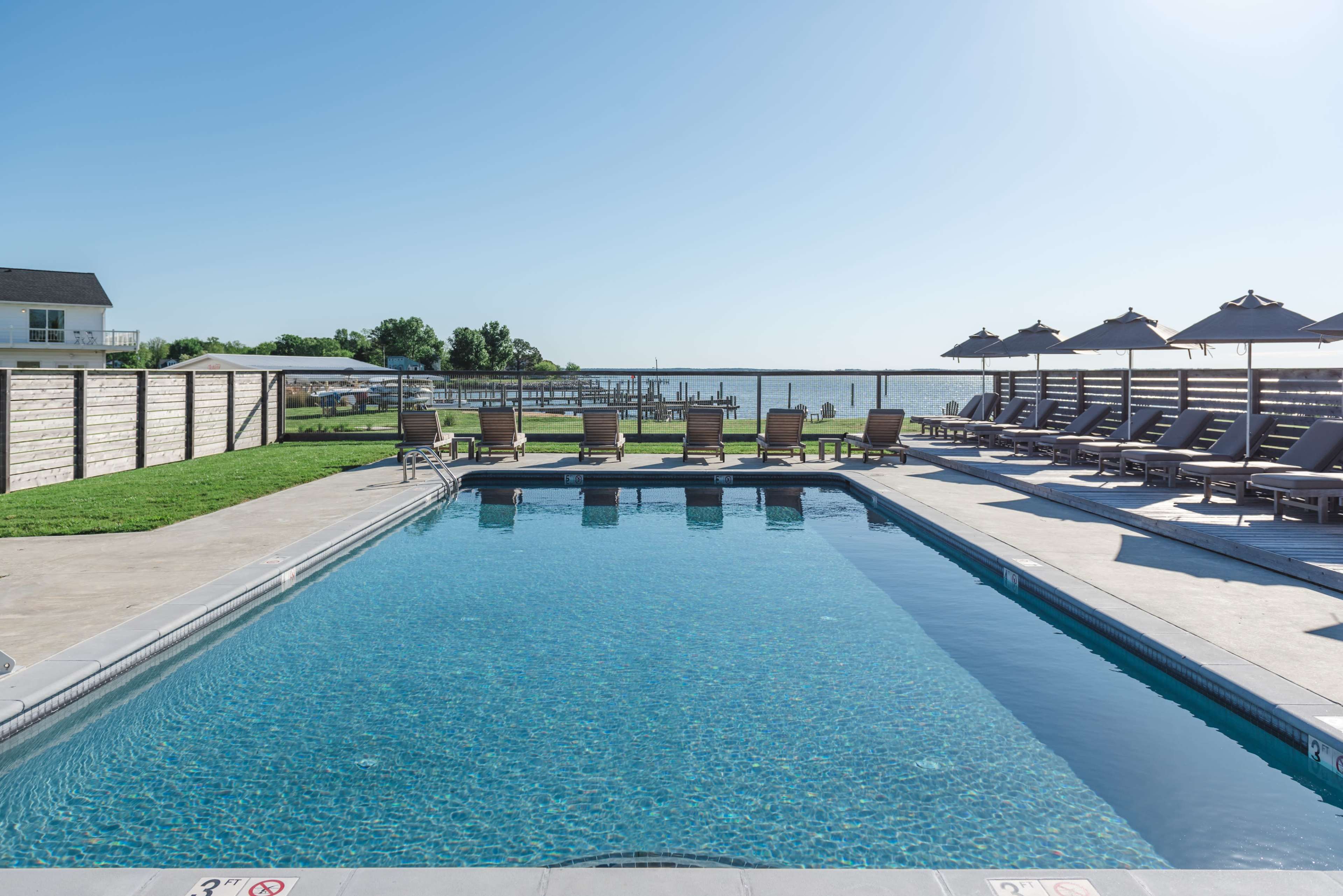 A clear swimming pool surrounded by lounge chairs and umbrellas, with a view of a waterfront in the background.