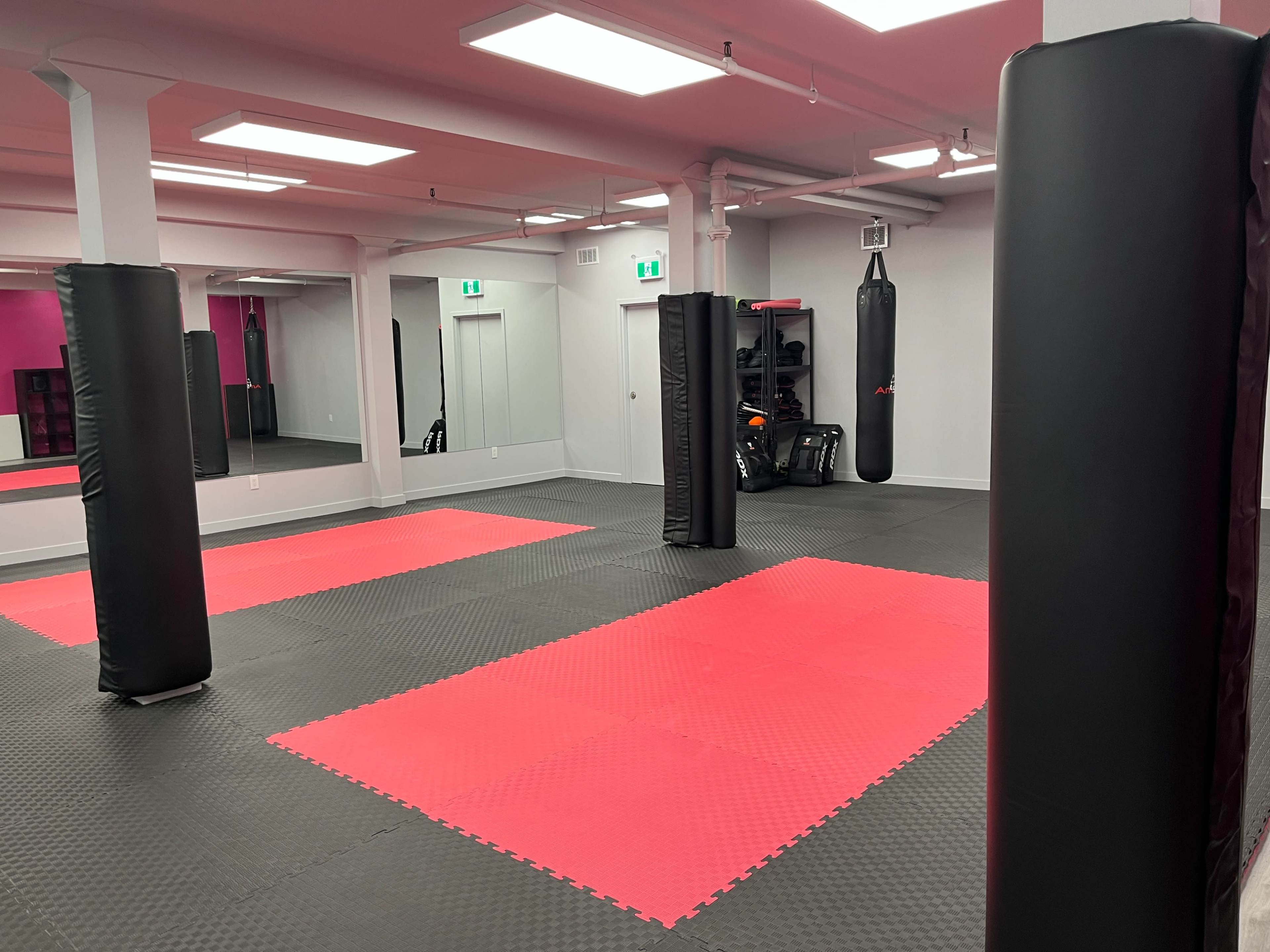 The image shows an empty gym interior designed for martial arts, featuring red and black mats, boxing bags, and protective pillars.