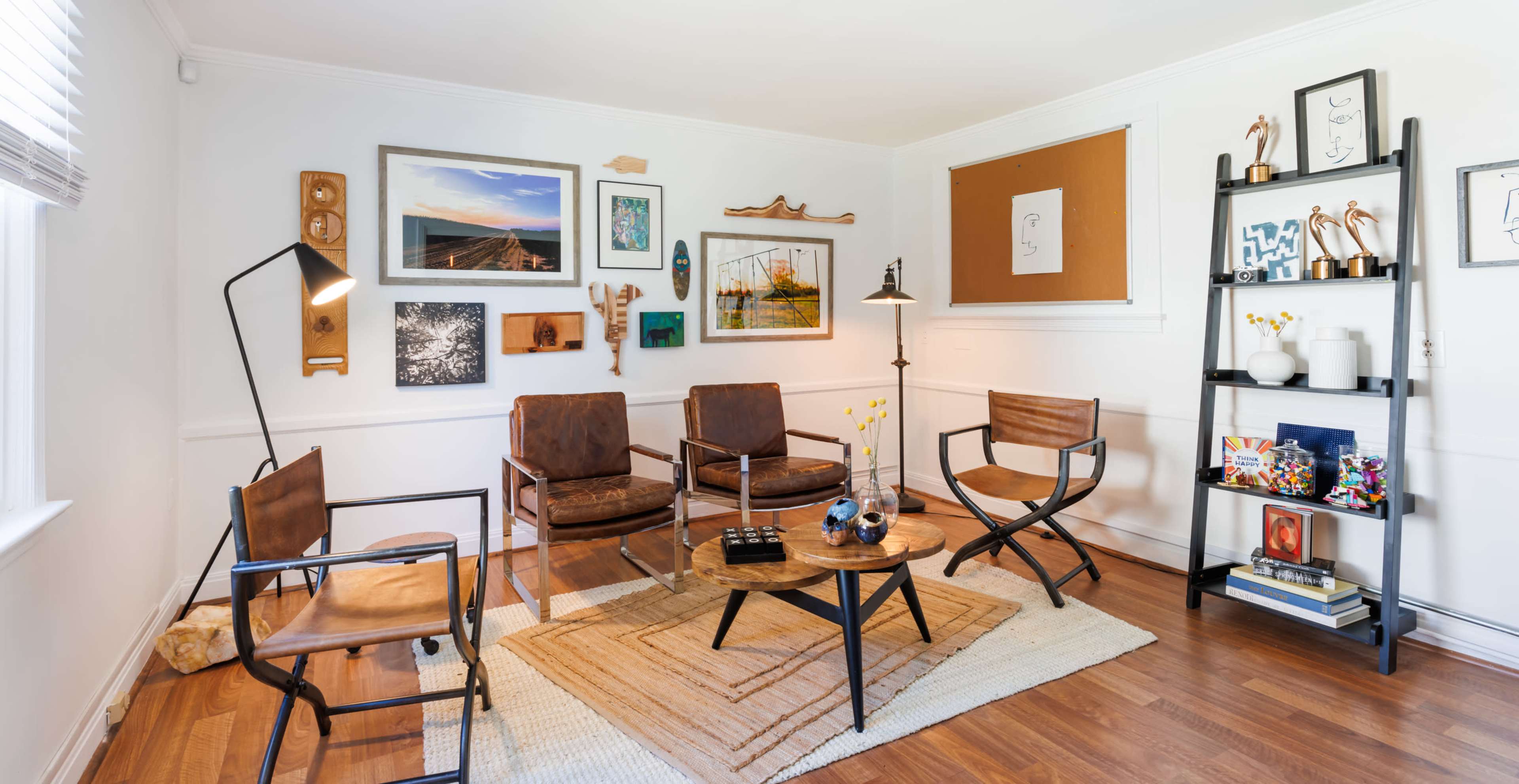 A cozy living room with four chairs arranged around a round coffee table on a woven rug, surrounded by wall art and a bookshelf filled with decorative items.