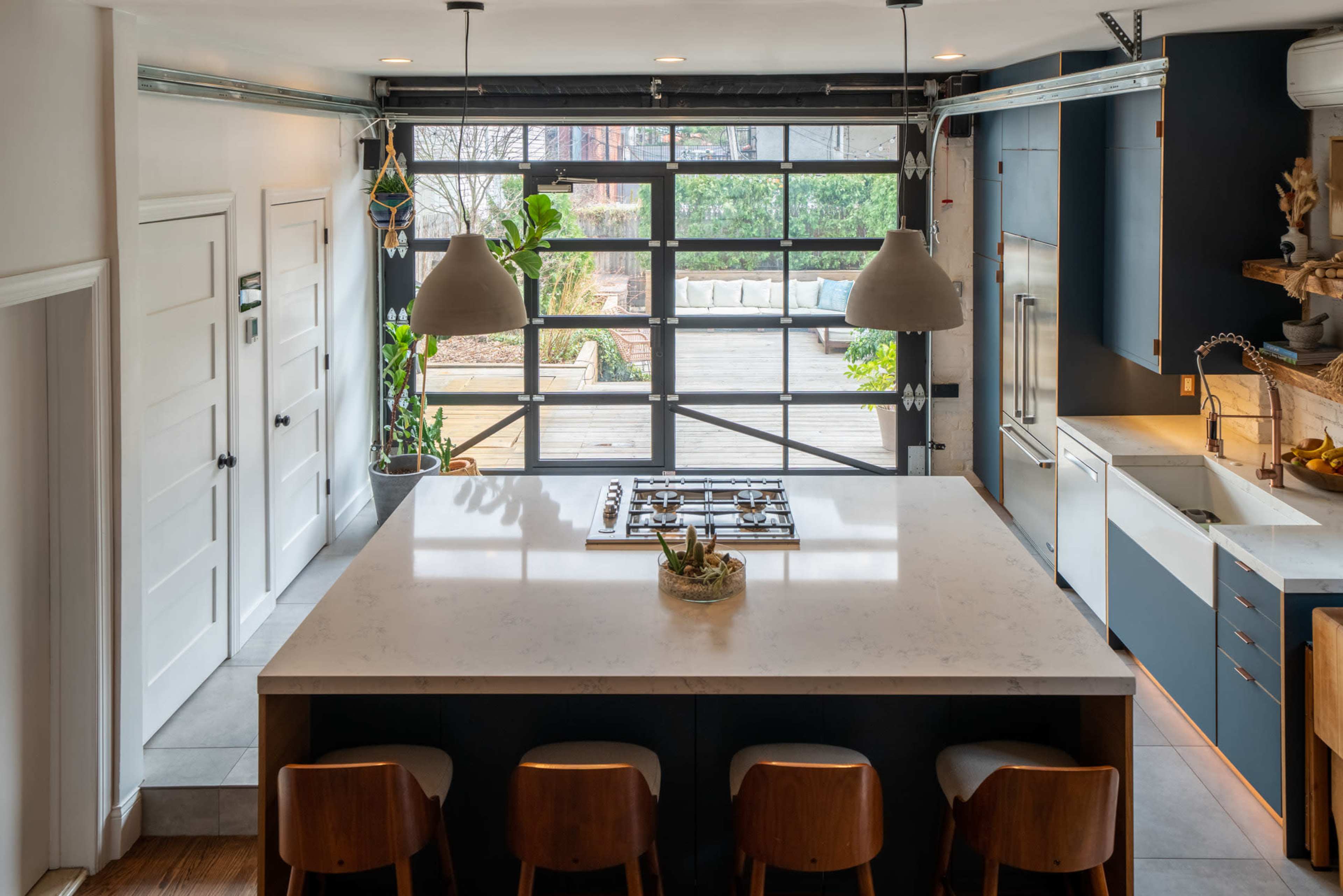 The image shows a modern kitchen featuring a large island with four wooden stools, a gas cooktop, and large windows overlooking an outdoor patio area.