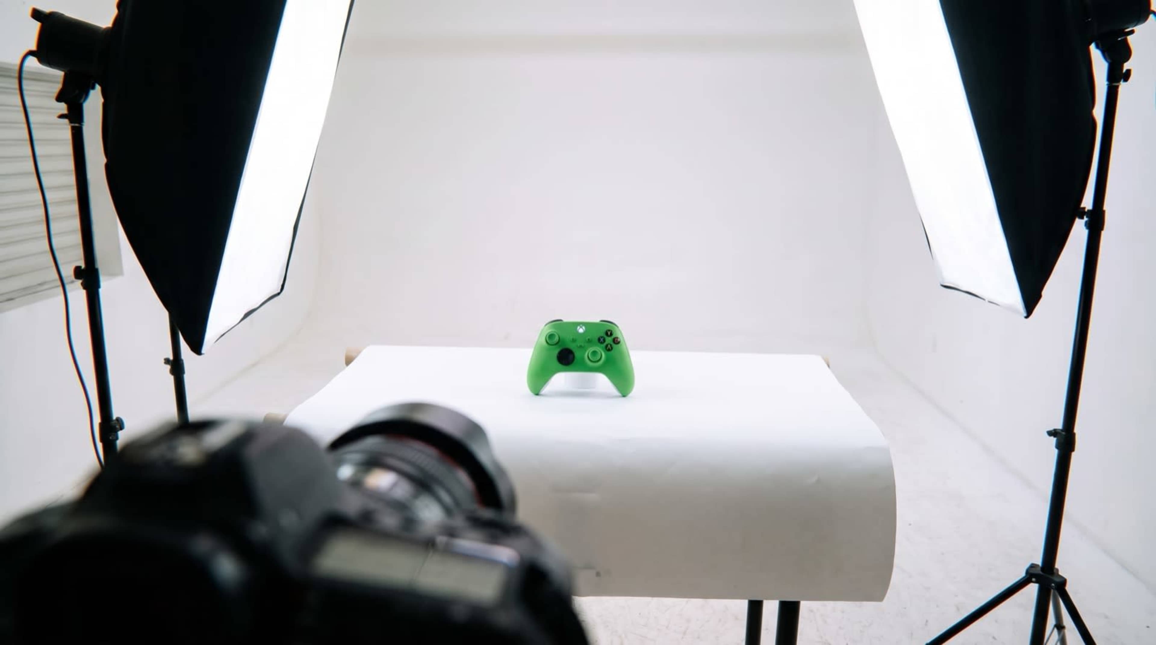 A green gaming controller is placed on a white table with softbox lights positioned on either side, and a camera is focused on it.