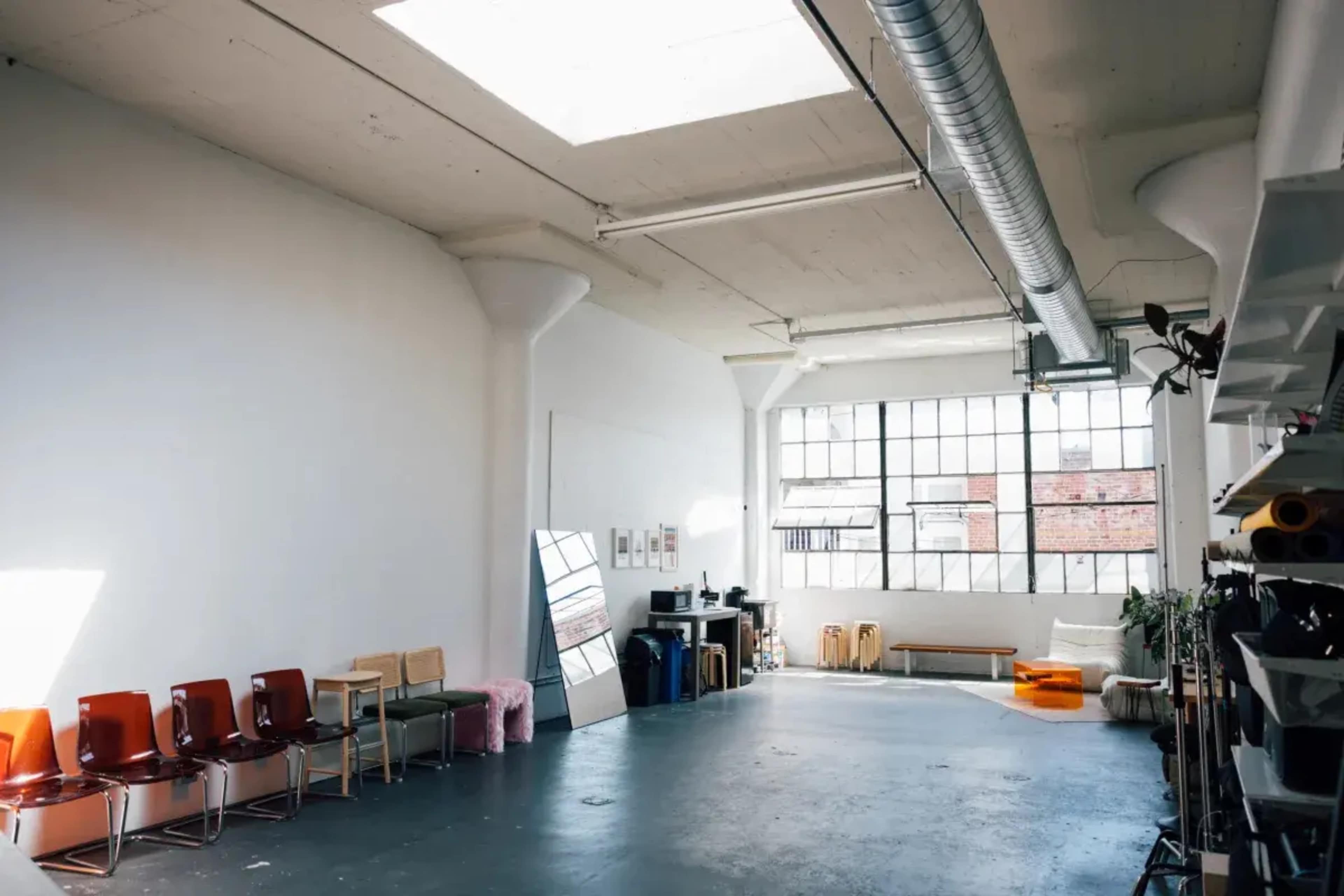 The image depicts a spacious, well-lit interior with a row of chairs along one wall and large windows allowing natural light to enter.