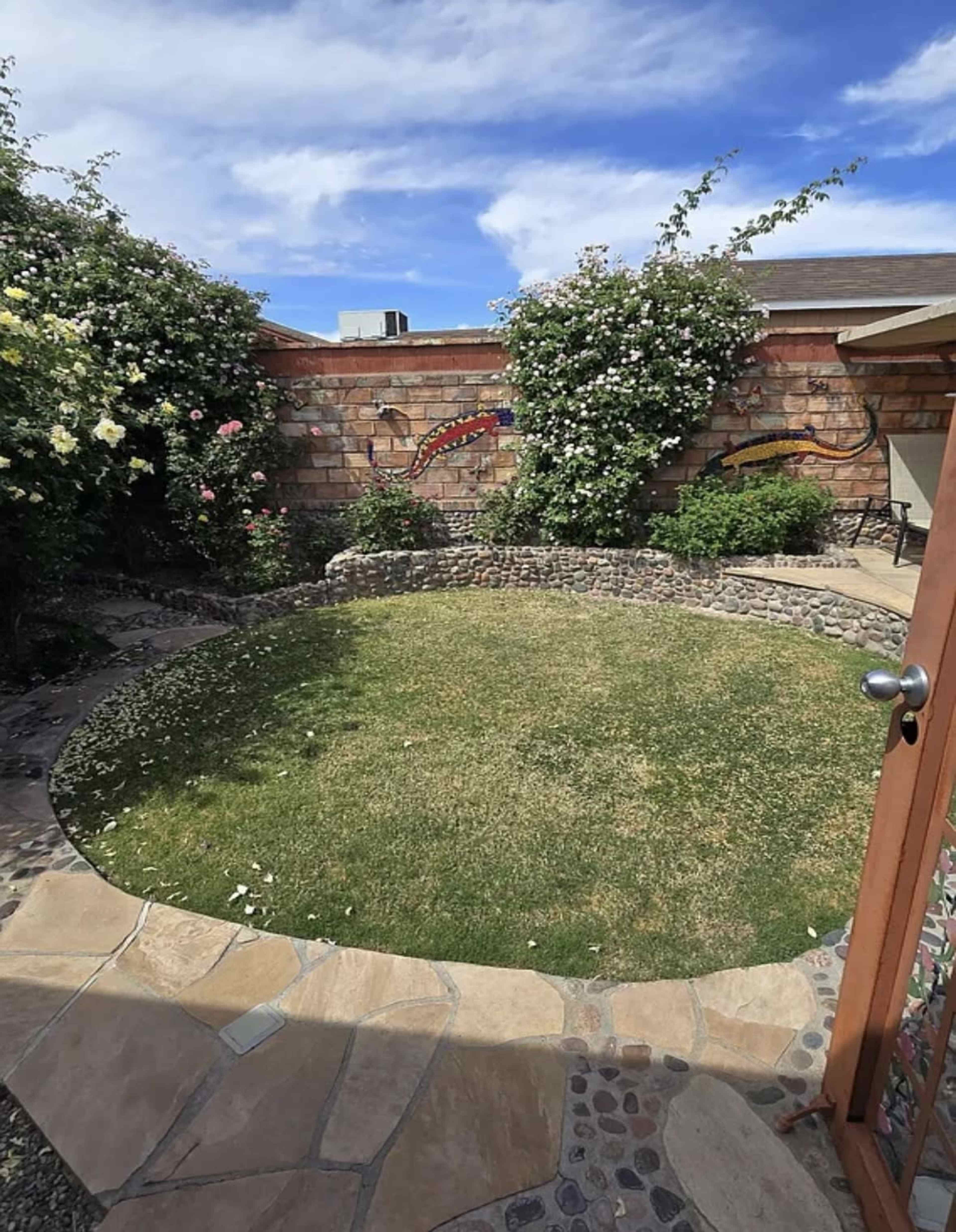 The image shows a circular patch of grass surrounded by a stone wall and flowering plants in a garden.