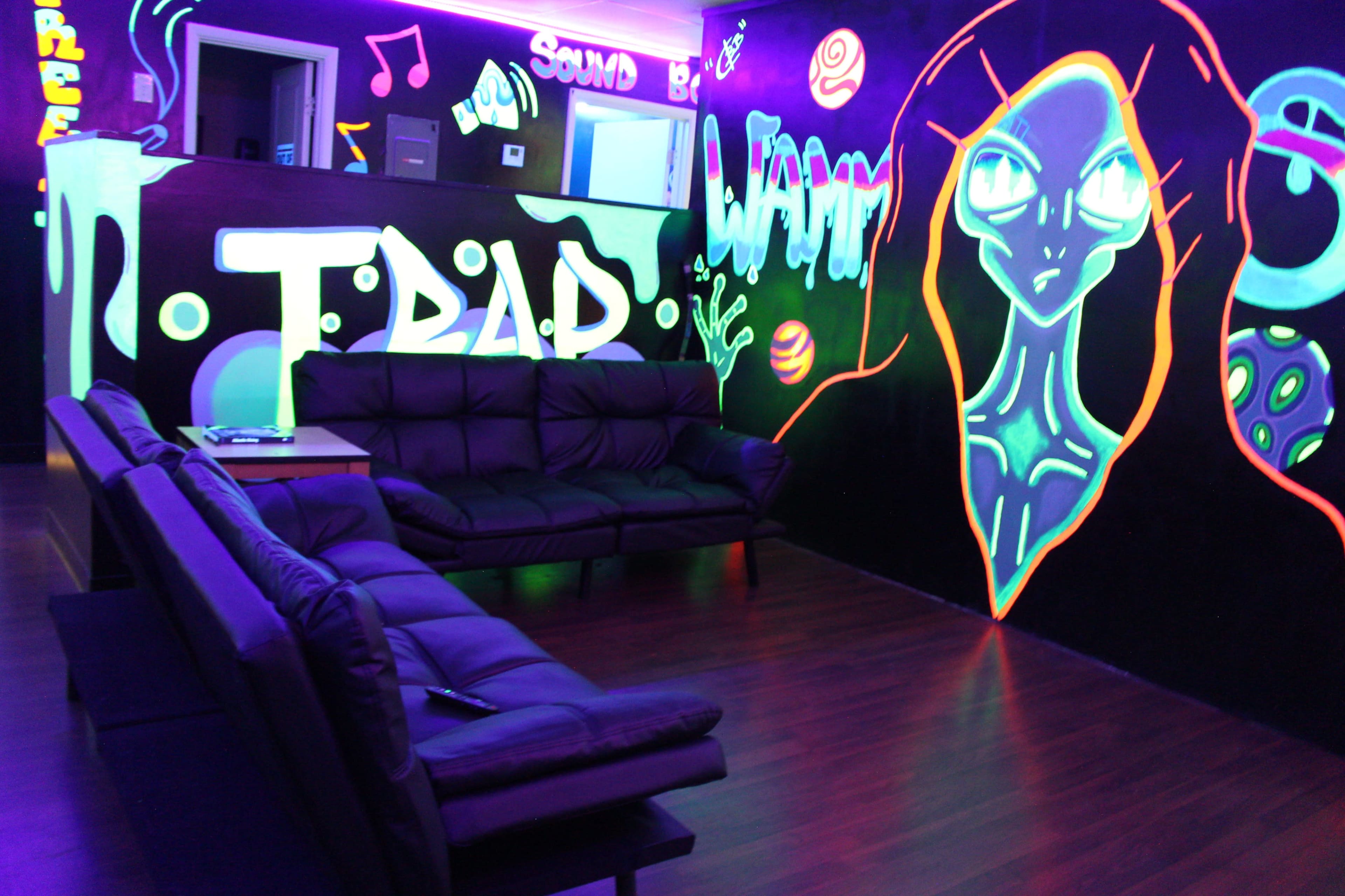 The image shows a lounge area with black walls decorated in neon graffiti, featuring a couch and vibrant lighting.