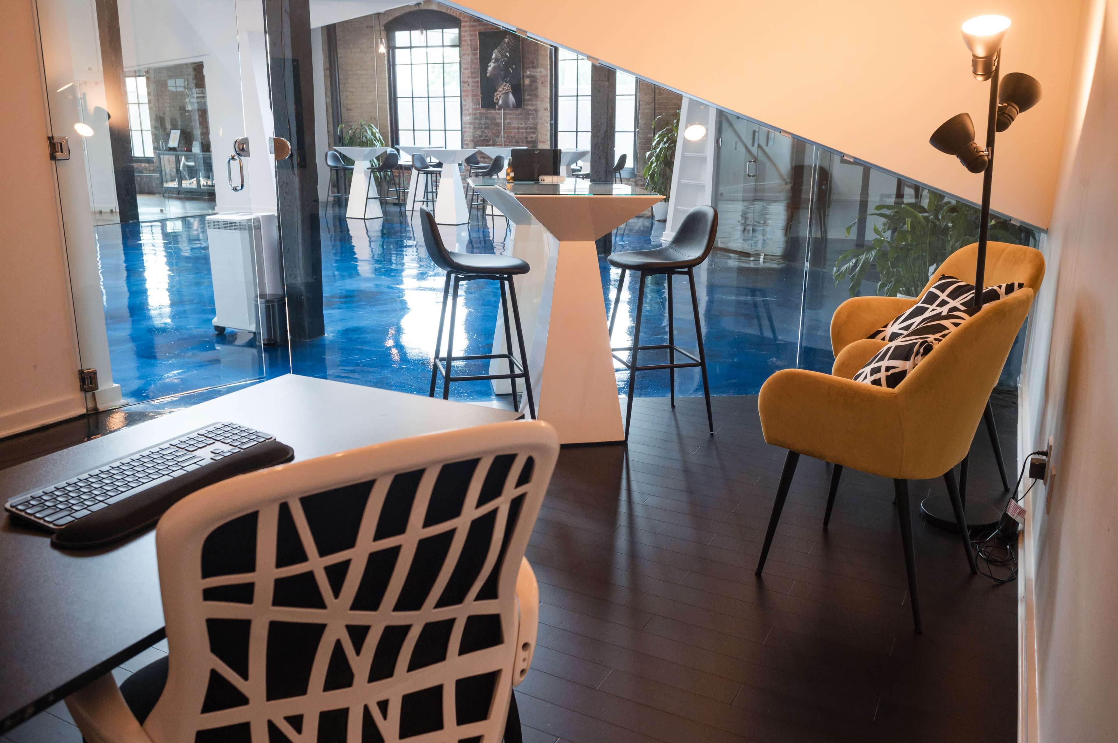A modern office space features a desk with a keyboard, a yellow chair, and a beverage table, with large windows illuminating the blue flooring.