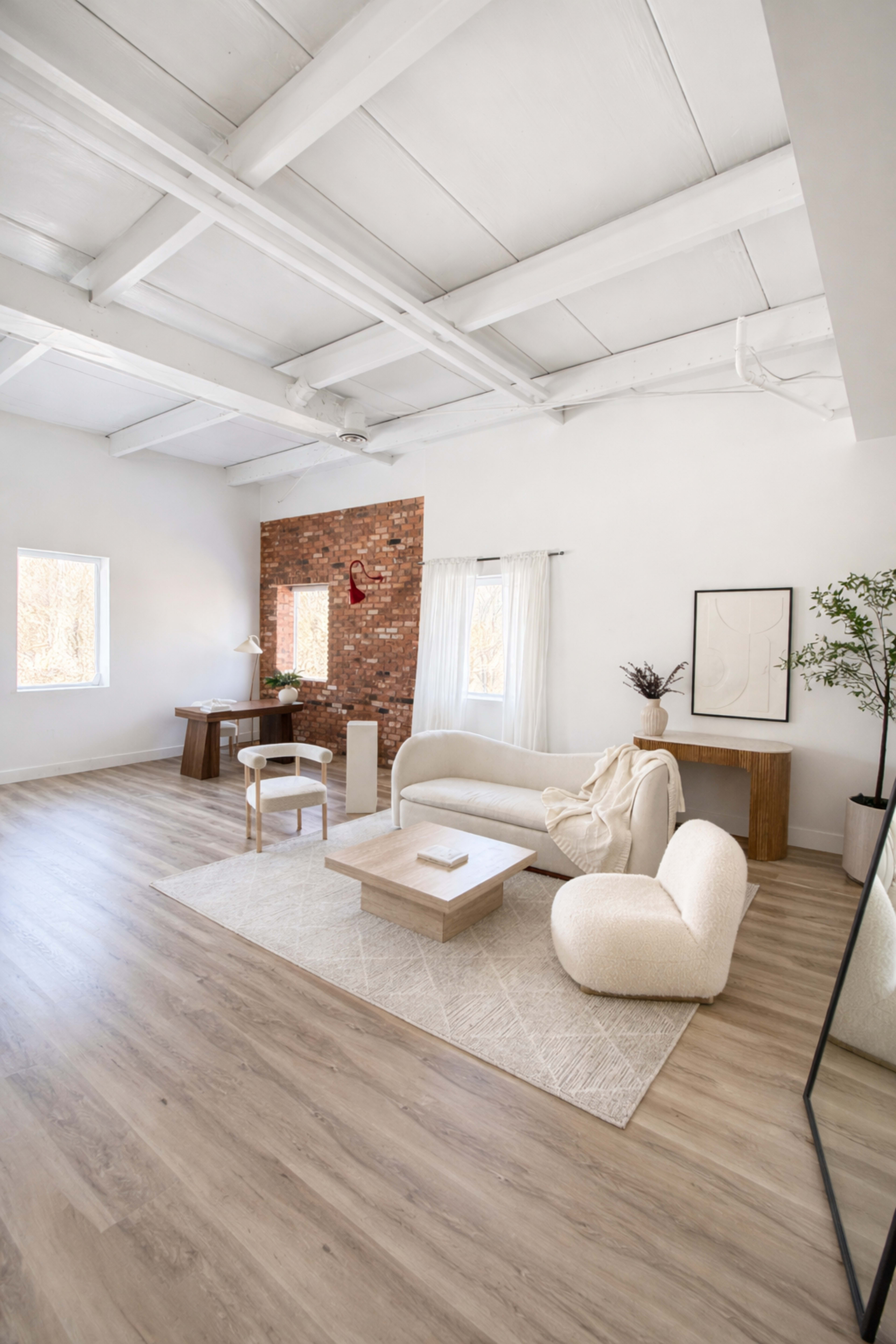 The image shows a bright, modern living room with a beige sectional sofa, a round chair, a wooden desk, and large windows illuminating the space.