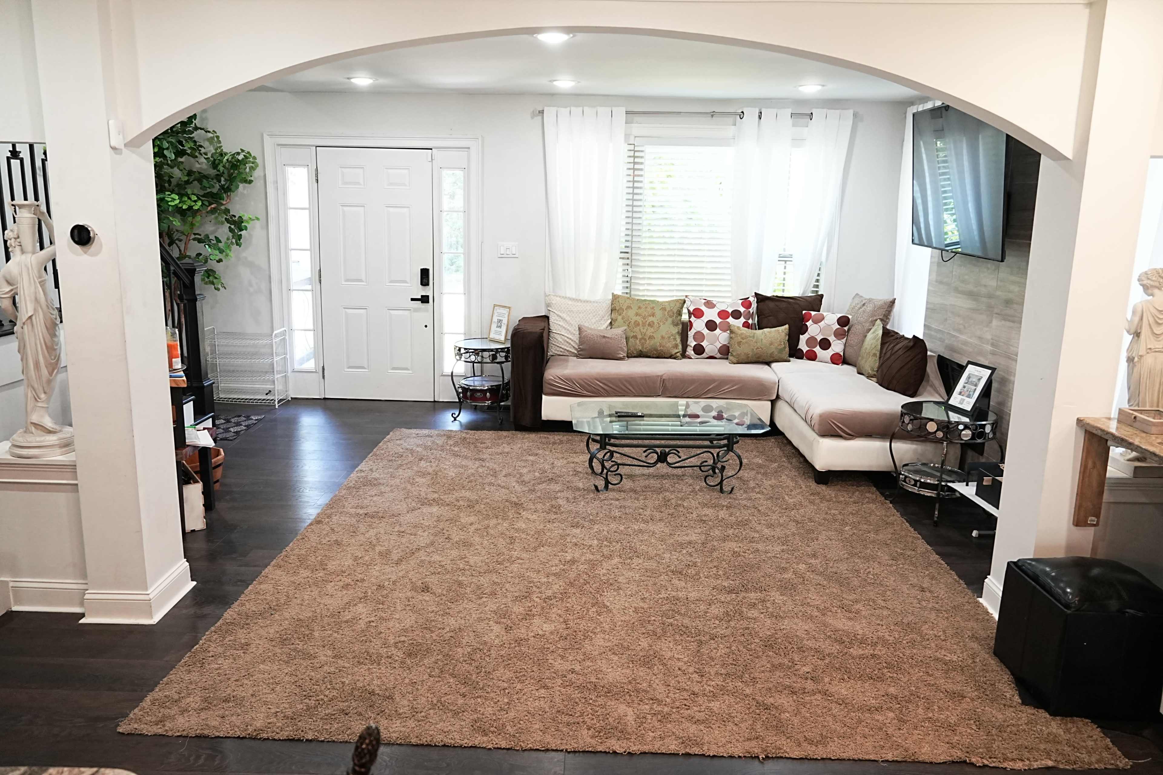 A living room with a sectional sofa adorned with colorful pillows, a glass coffee table, and a large area rug on a dark wooden floor.