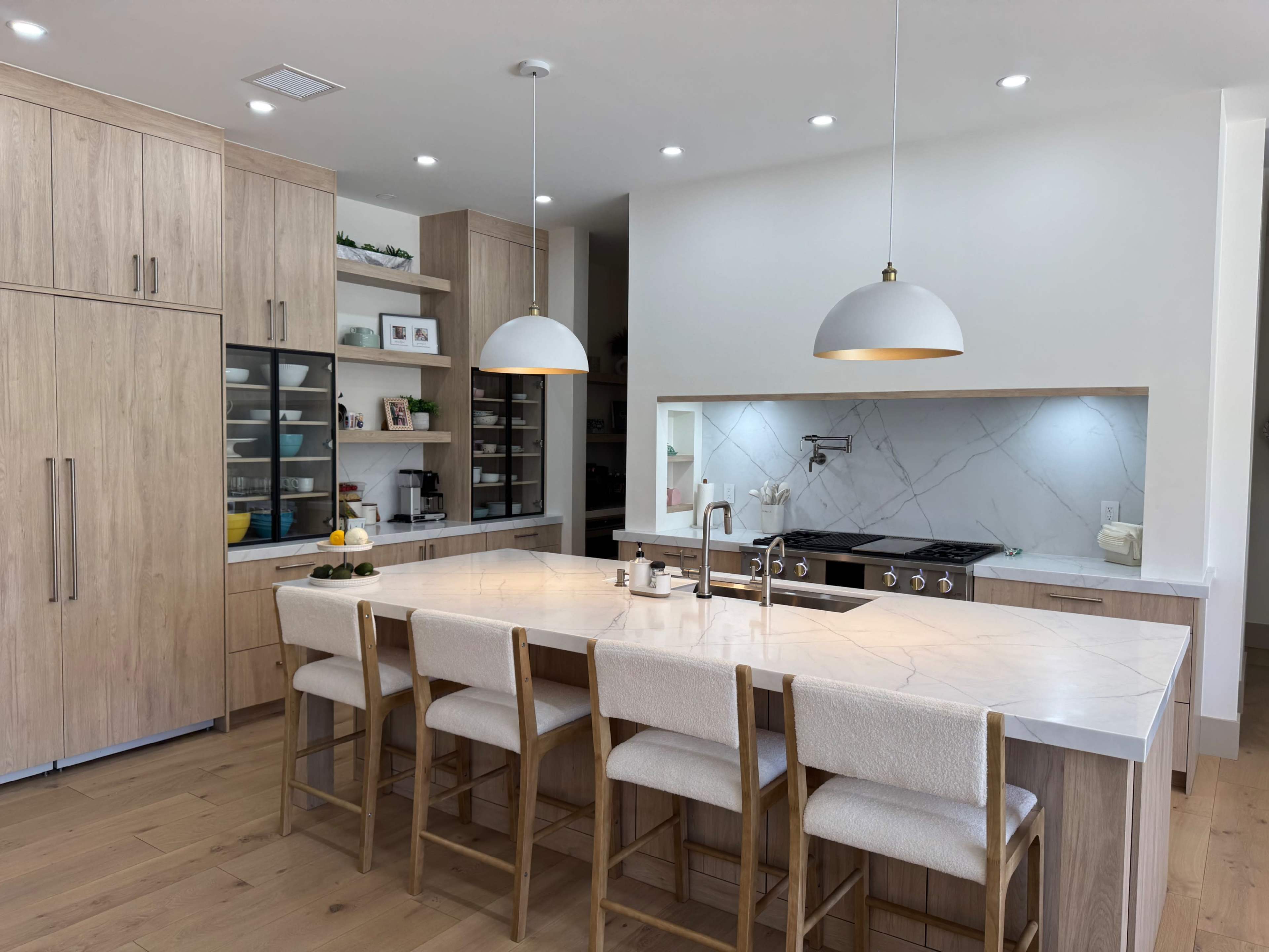 The image shows a modern kitchen with light wood cabinetry, a large island with seating, and a marble countertop.
