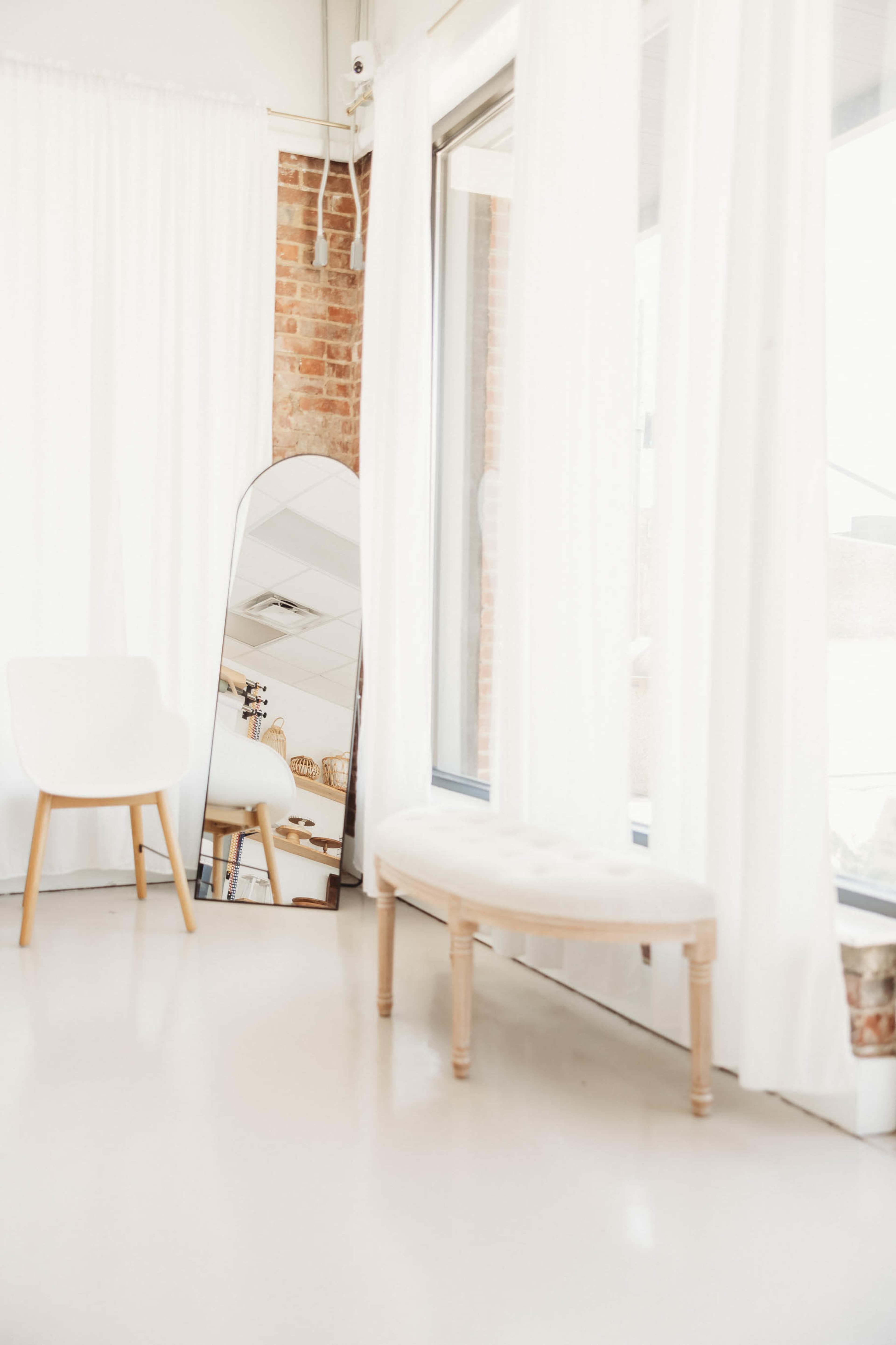 The image shows a bright interior space with white curtains, a brick wall, a sleek chair, a bench, and a tall mirror.