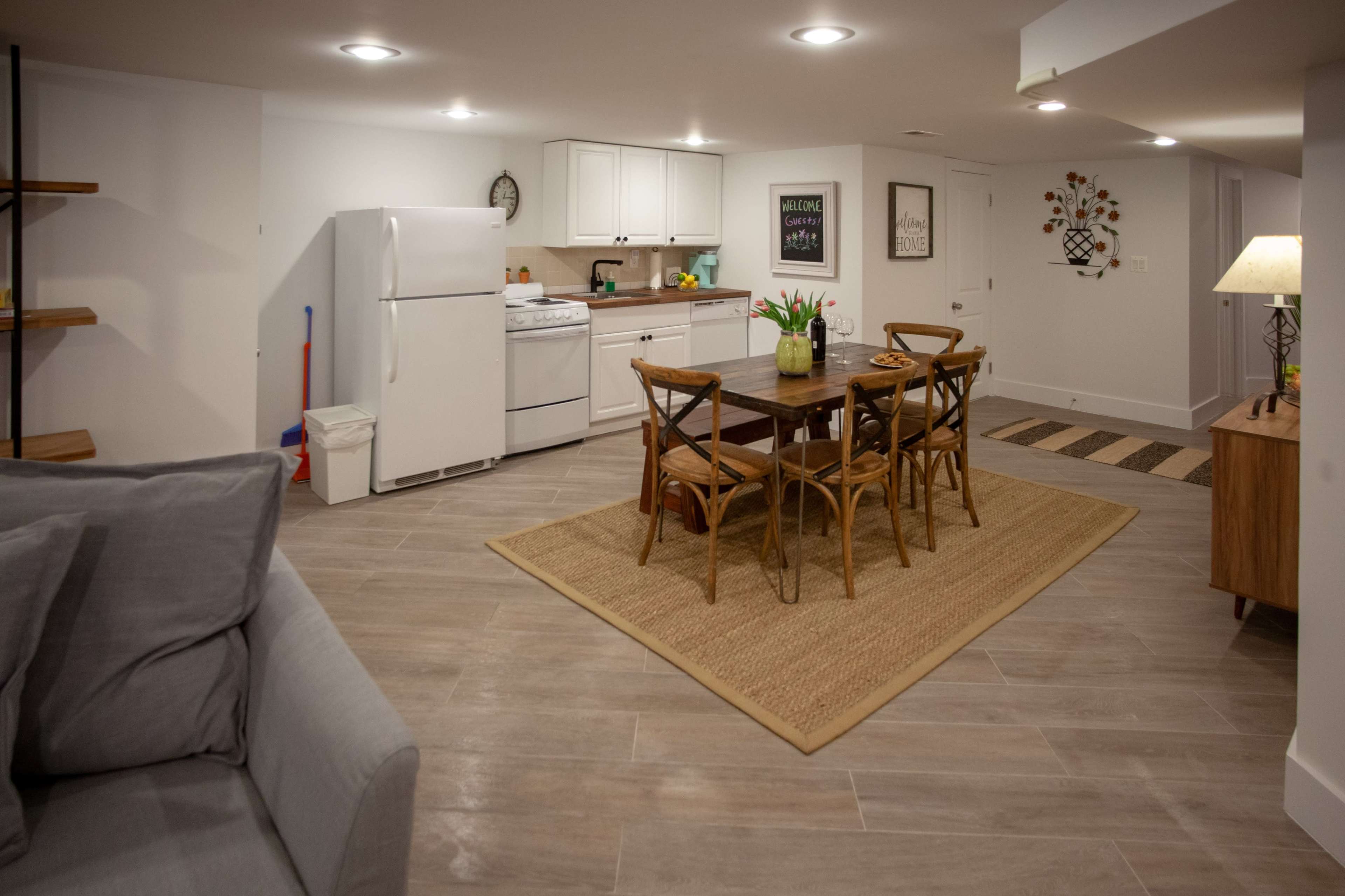 A modern basement space featuring a kitchen area with white appliances, a dining table with four chairs, and a cozy seating area with a gray sofa.
