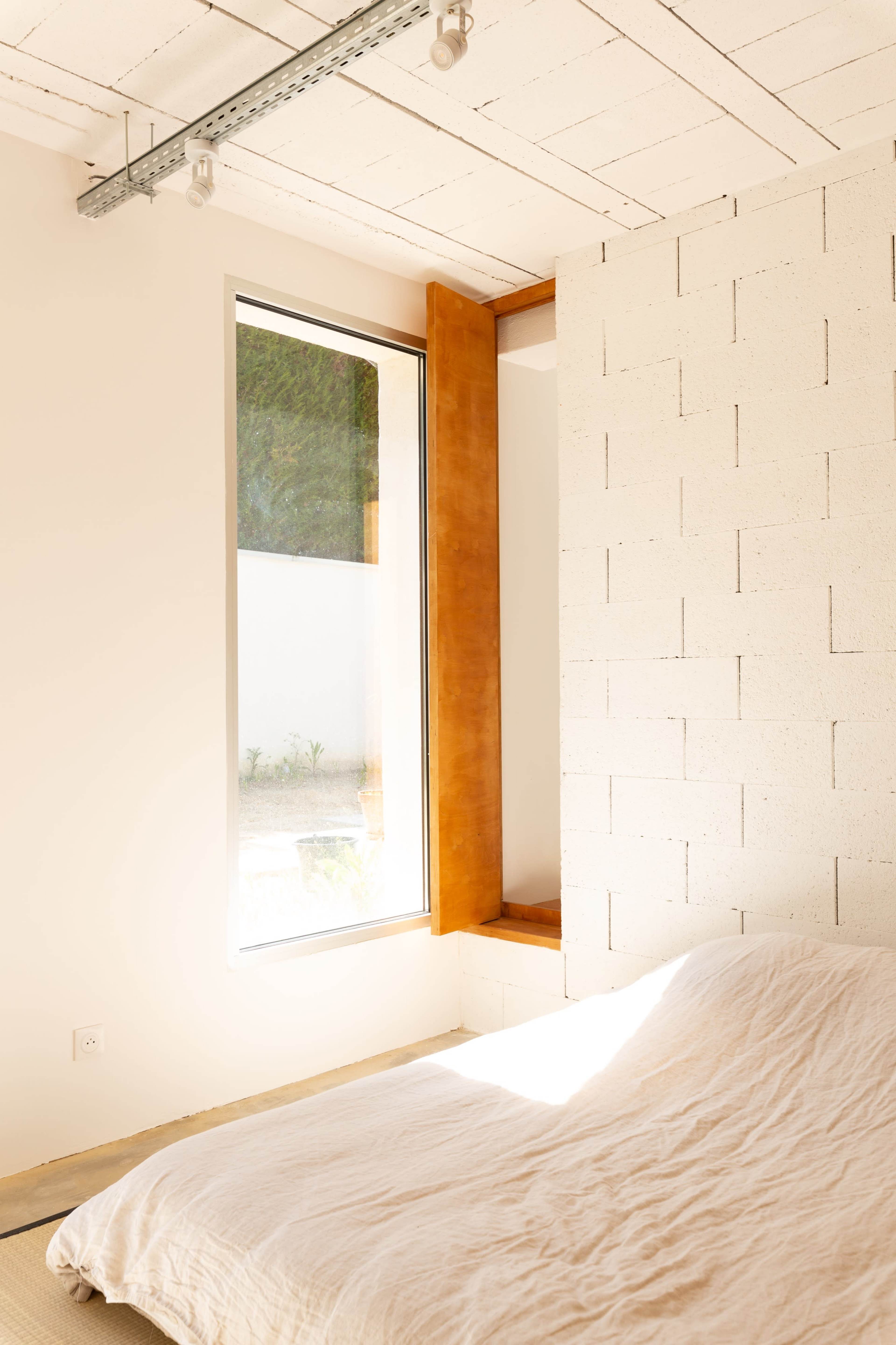 A minimal bedroom features a bed with a white coverlet, a large window with wooden trim, and a patterned concrete wall.