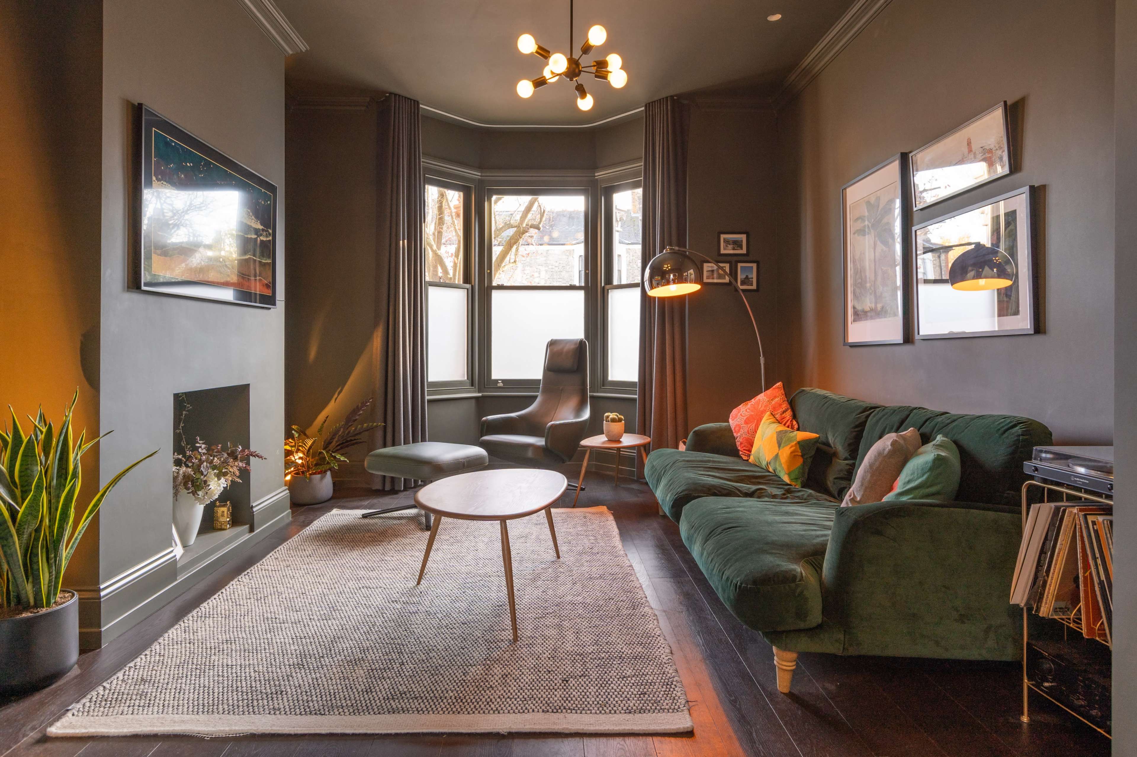 The image shows a cozy living room with a green velvet sofa, a round wooden coffee table, and a modern floor lamp, all set against dark walls and large windows.