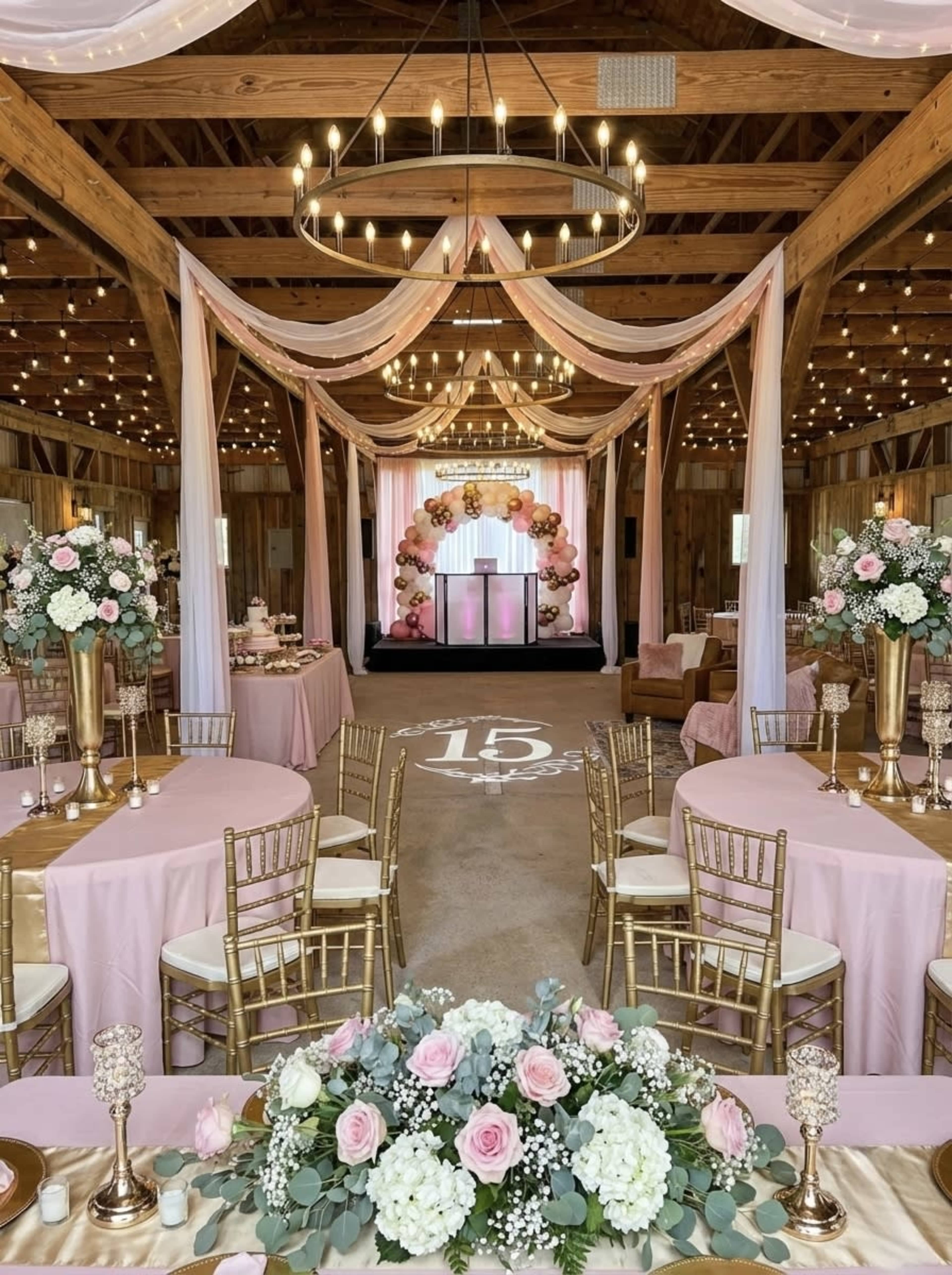 The image shows a decorated indoor venue featuring elegant tables with pink tablecloths, floral centerpieces, and a backdrop of draped fabric and balloons, illuminated by string lights.