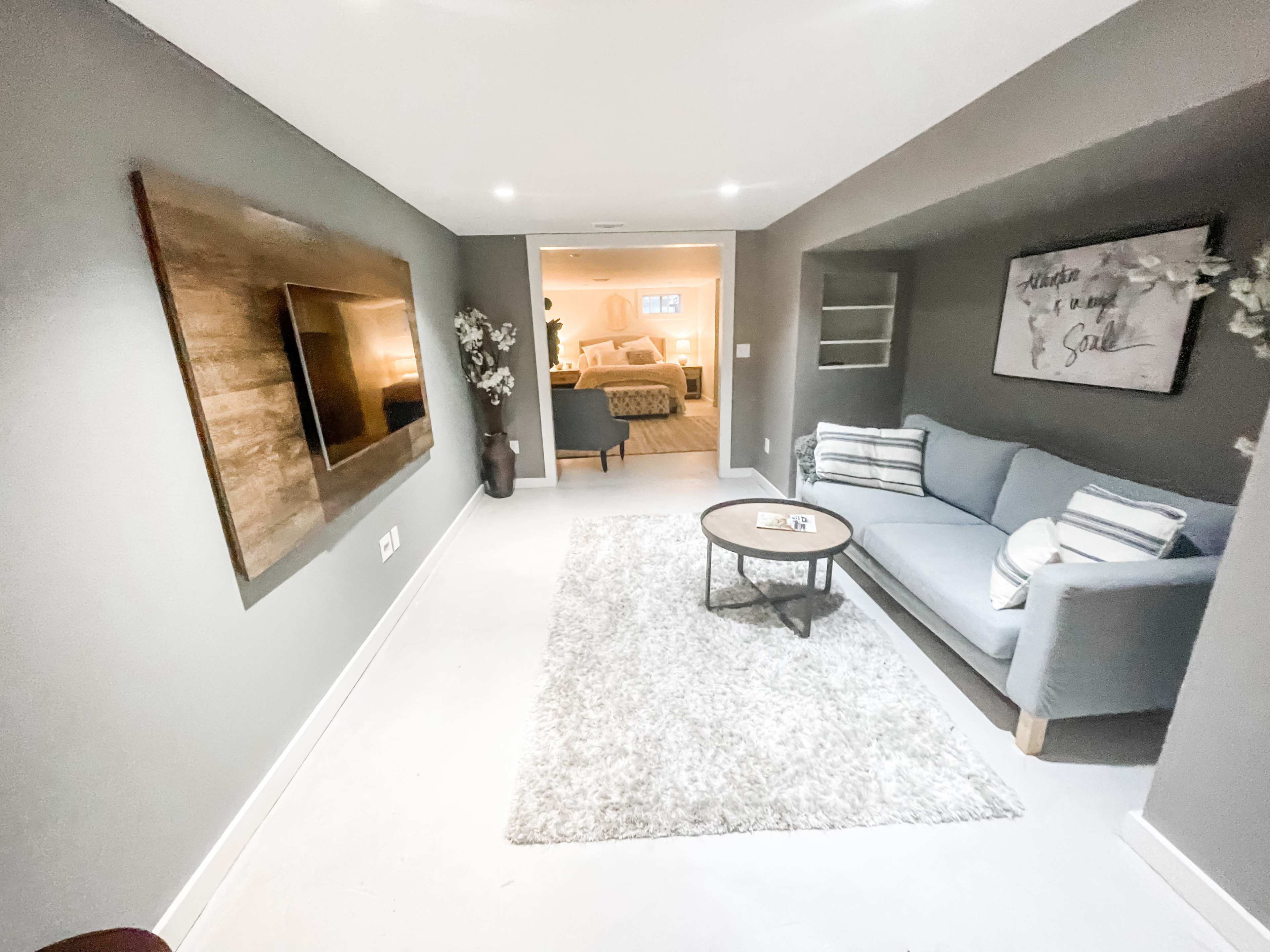 A modern basement living area features a gray sofa, a round table, and a wall-mounted TV, with light streaming in from an open doorway.